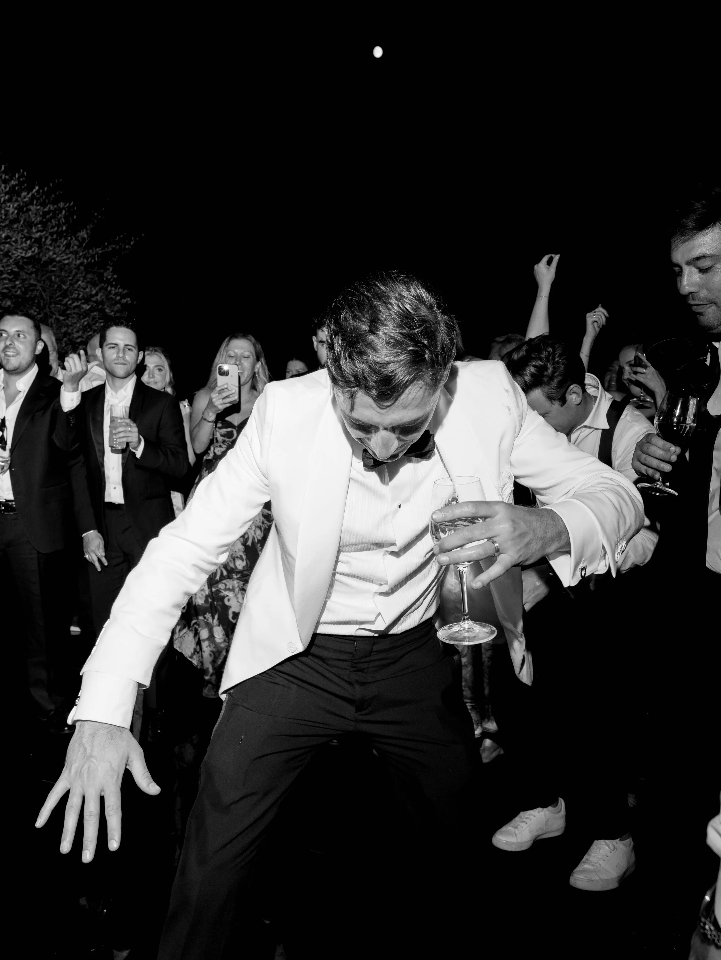 A black and white photo of a lively party with guests dancing and enjoying drinks, with a man in a tuxedo in the foreground holding a glass of wine and others around him taking photos or dancing.