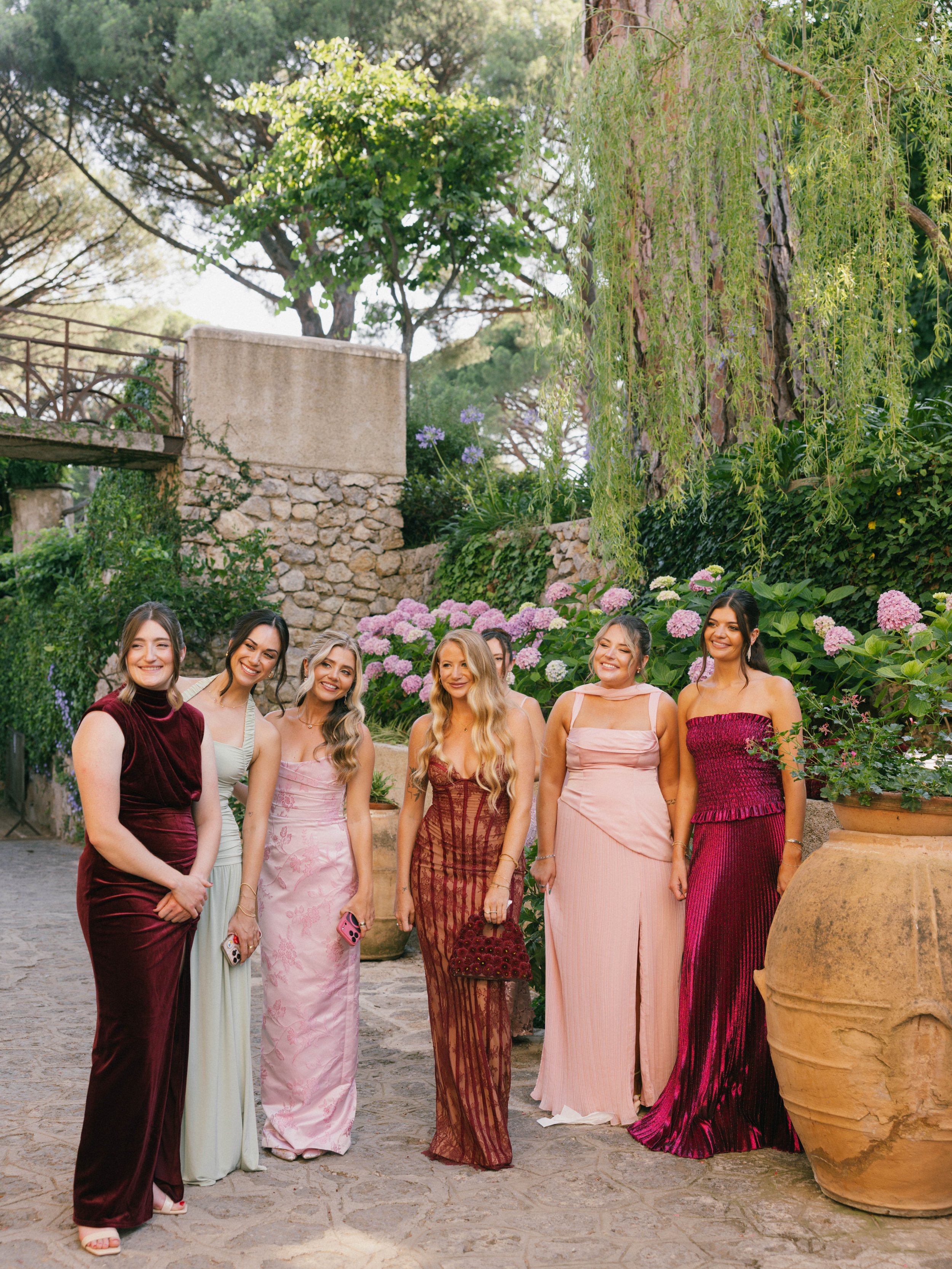 Group of women in colorful formal dresses standing outdoors in a garden with stone walls, large pottery, and lush greenery.