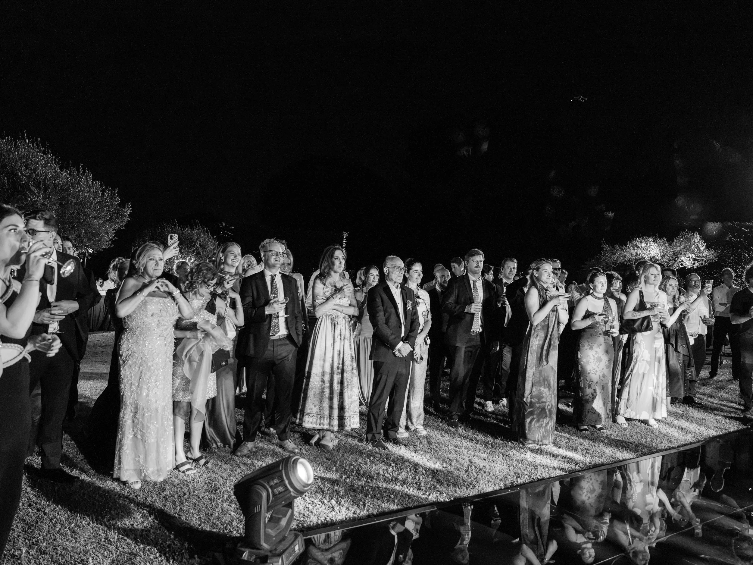 People dressed in formal attire attending an outdoor night event, standing on grass with trees and strategic lighting.