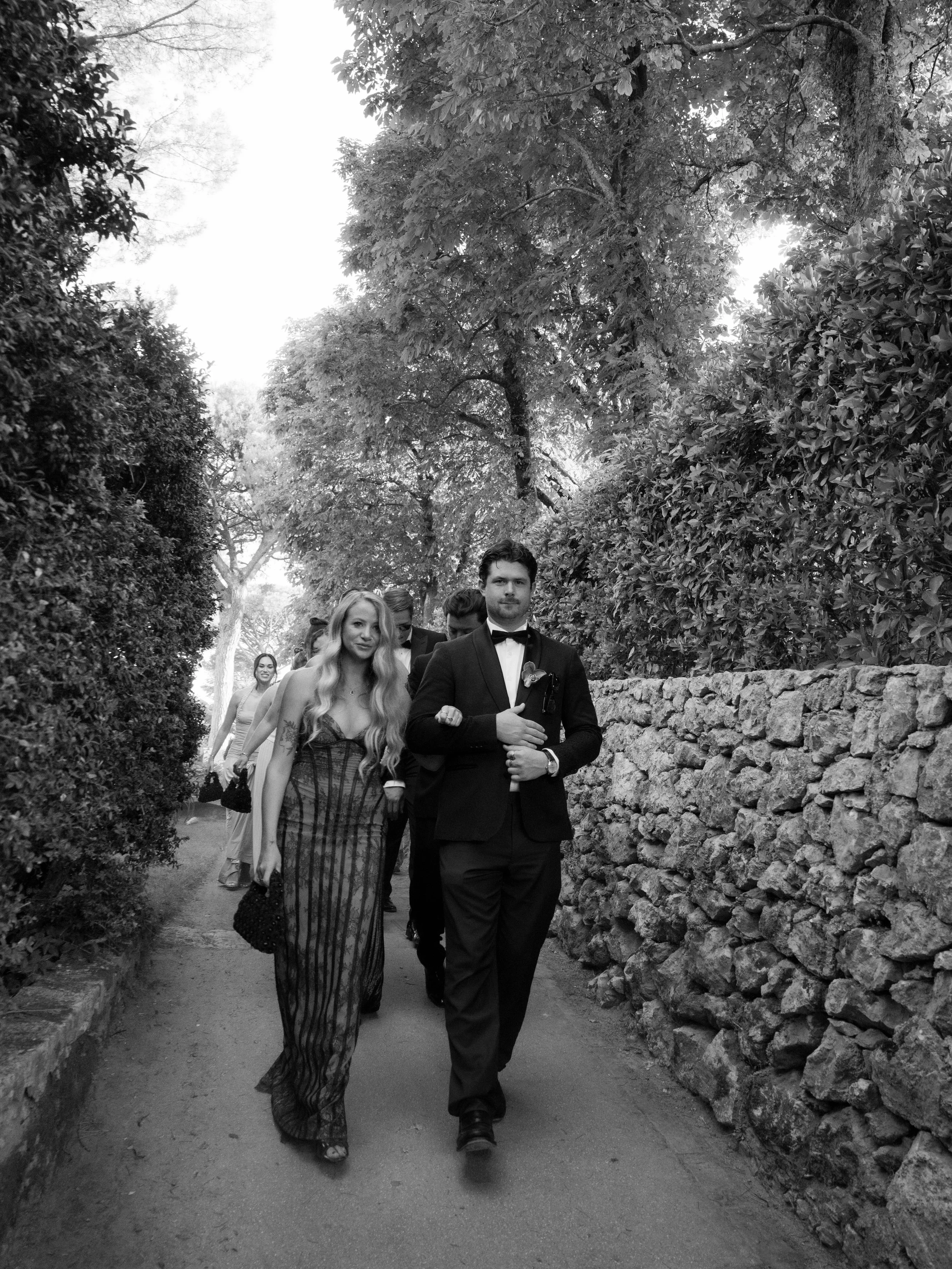 A group of people dressed in formal clothing walking along a narrow path lined with trees and a stone wall on one side. Villa Cimbrone wedding Amalfi Coast