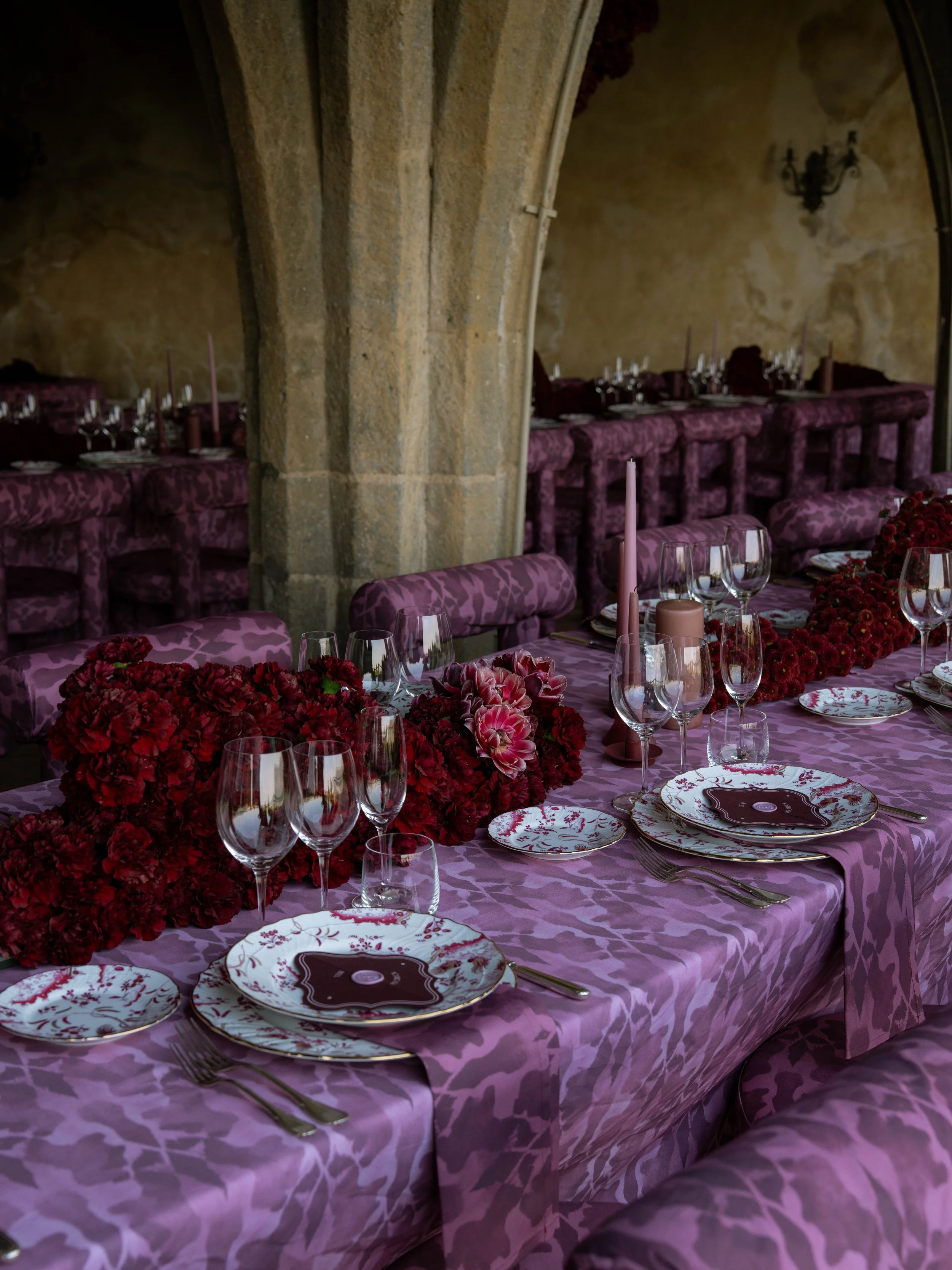 A decorated dining table with purple floral patterned tablecloth, red floral centerpieces, pink candles, fine china with floral design, and empty wine glasses. The background features stone archways and purple upholstered chairs.