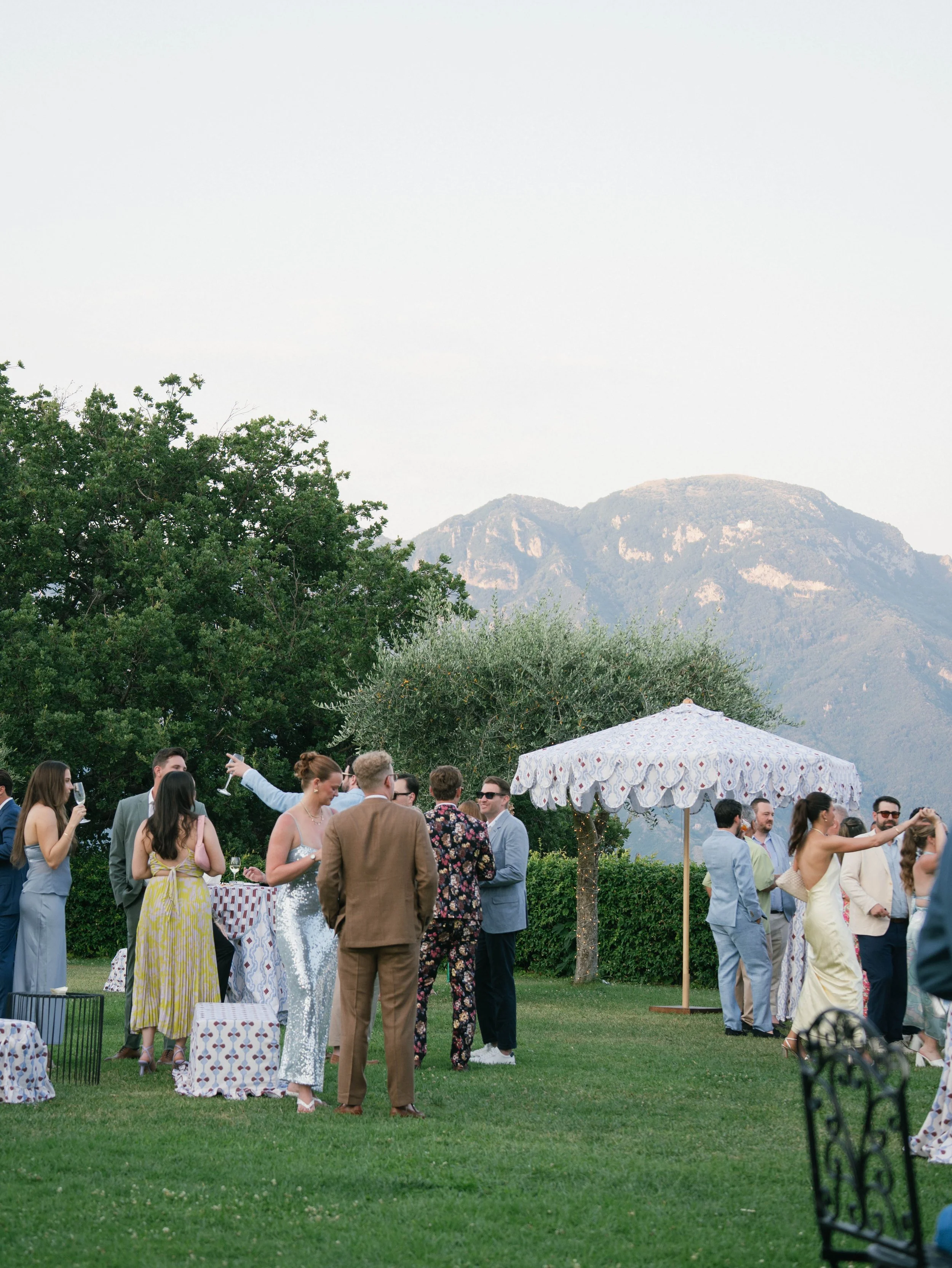 People gathered outdoors at an event under a large umbrella with a mountainous landscape in the background.