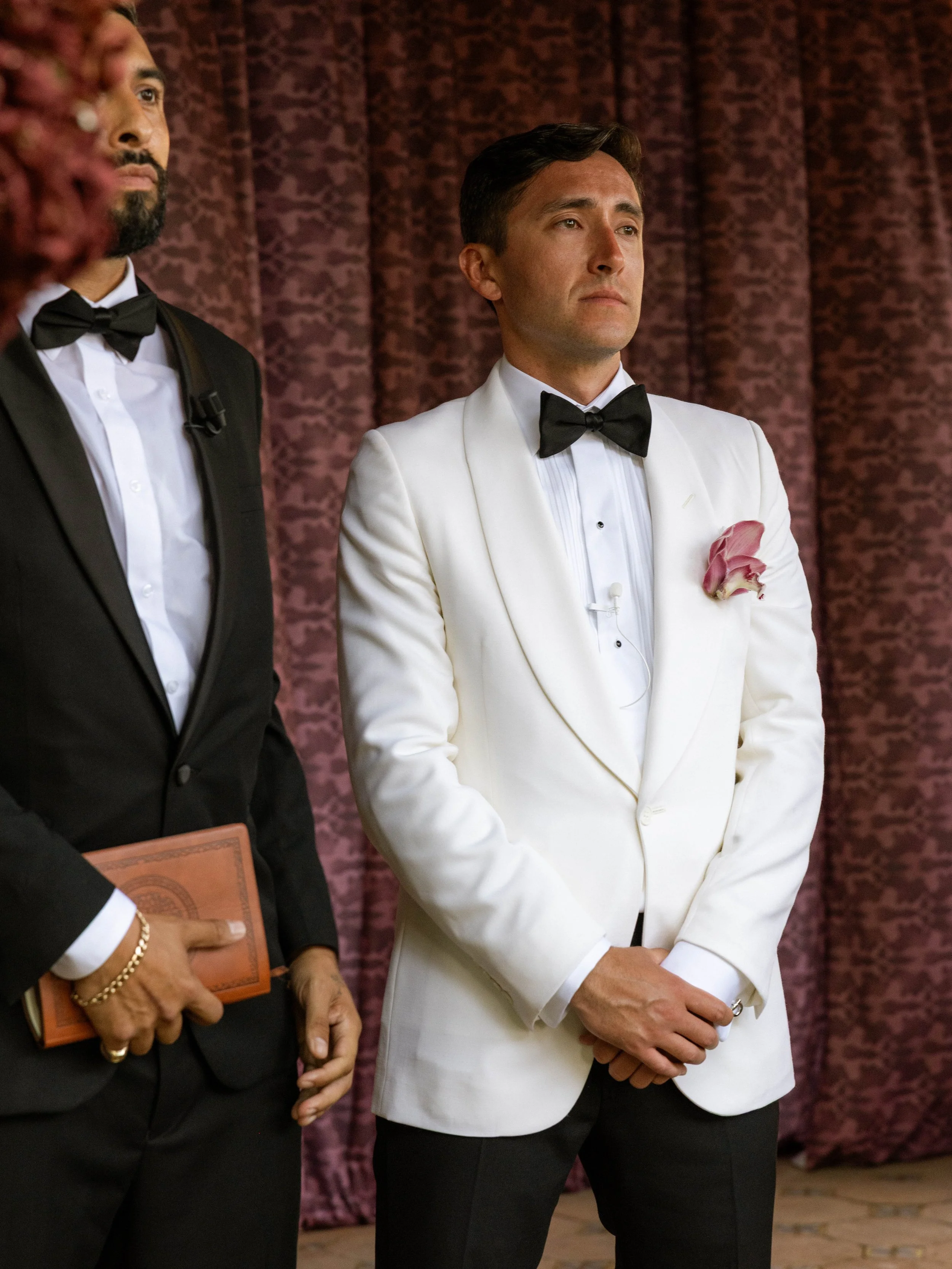 Two men dressed in tuxedos standing side by side, with the man on the right wearing a white tuxedo jacket and black bow tie, against a maroon patterned curtain background.
