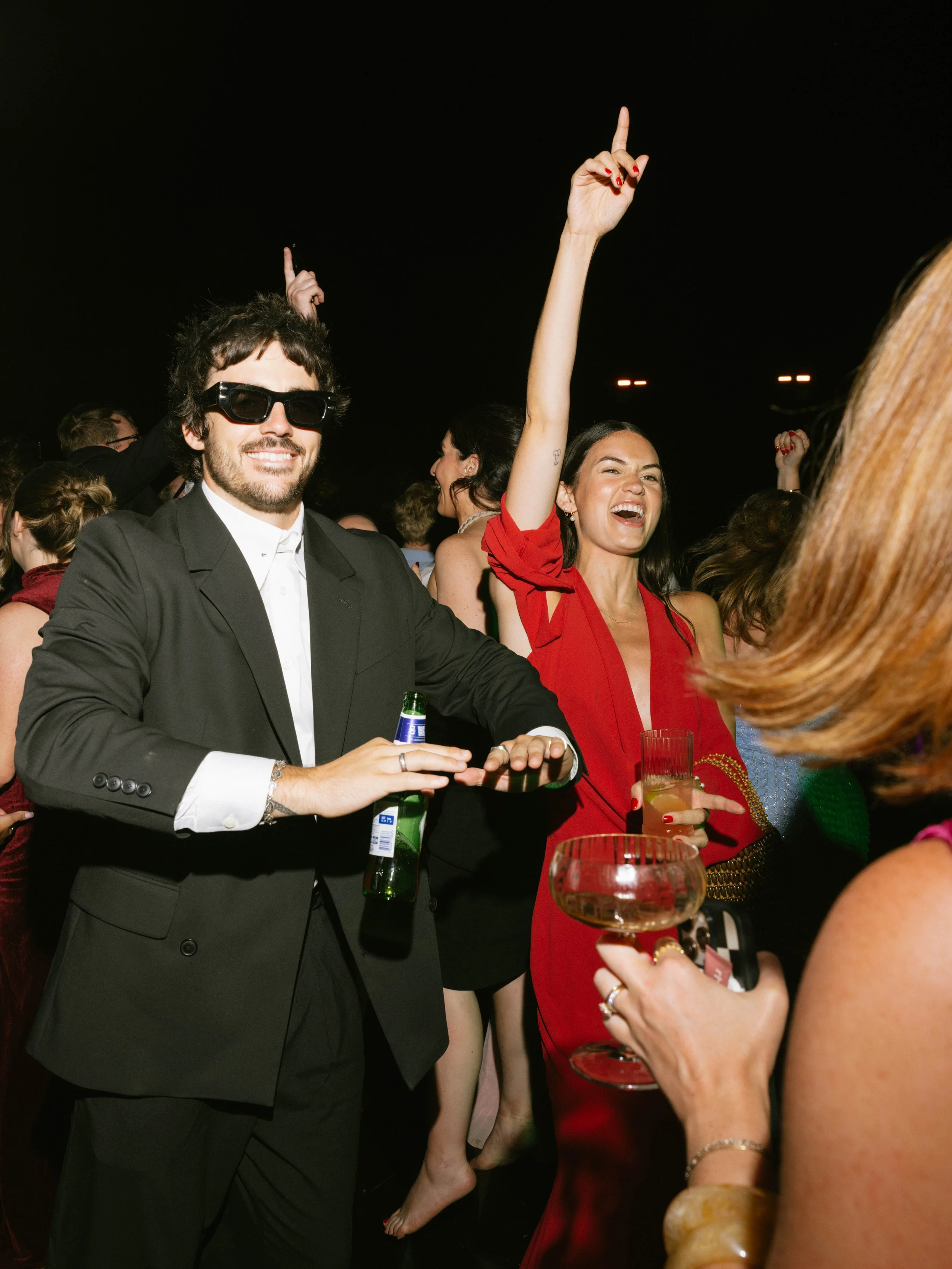 People dancing and celebrating at a party or nightclub, smiling, raising drinks and hands, dressed in elegant attire, nighttime scene.