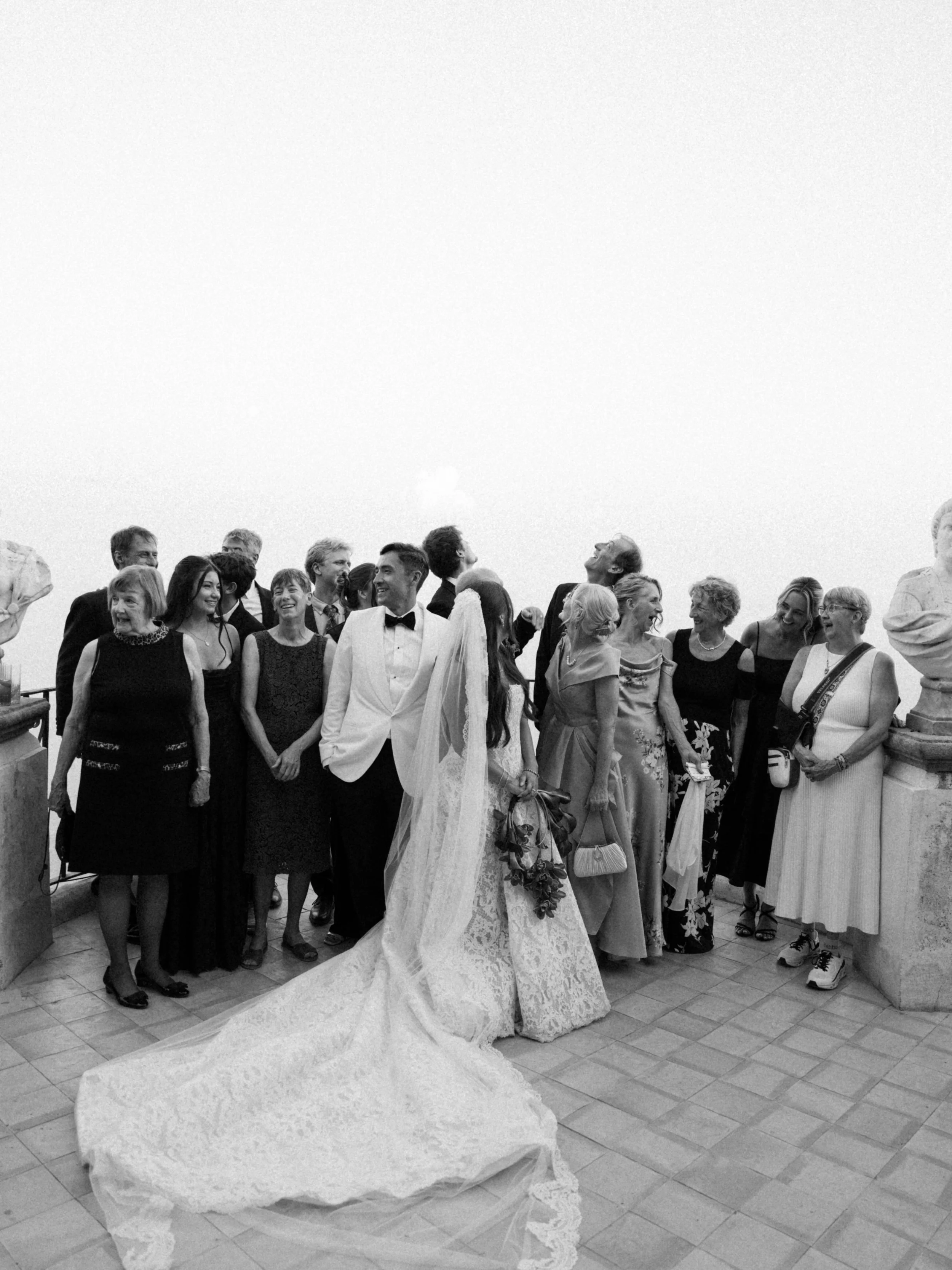 A bride in a wedding dress and veil with a long train, talking to a group of people, mostly women, dressed in formal attire, at a wedding reception or celebration.