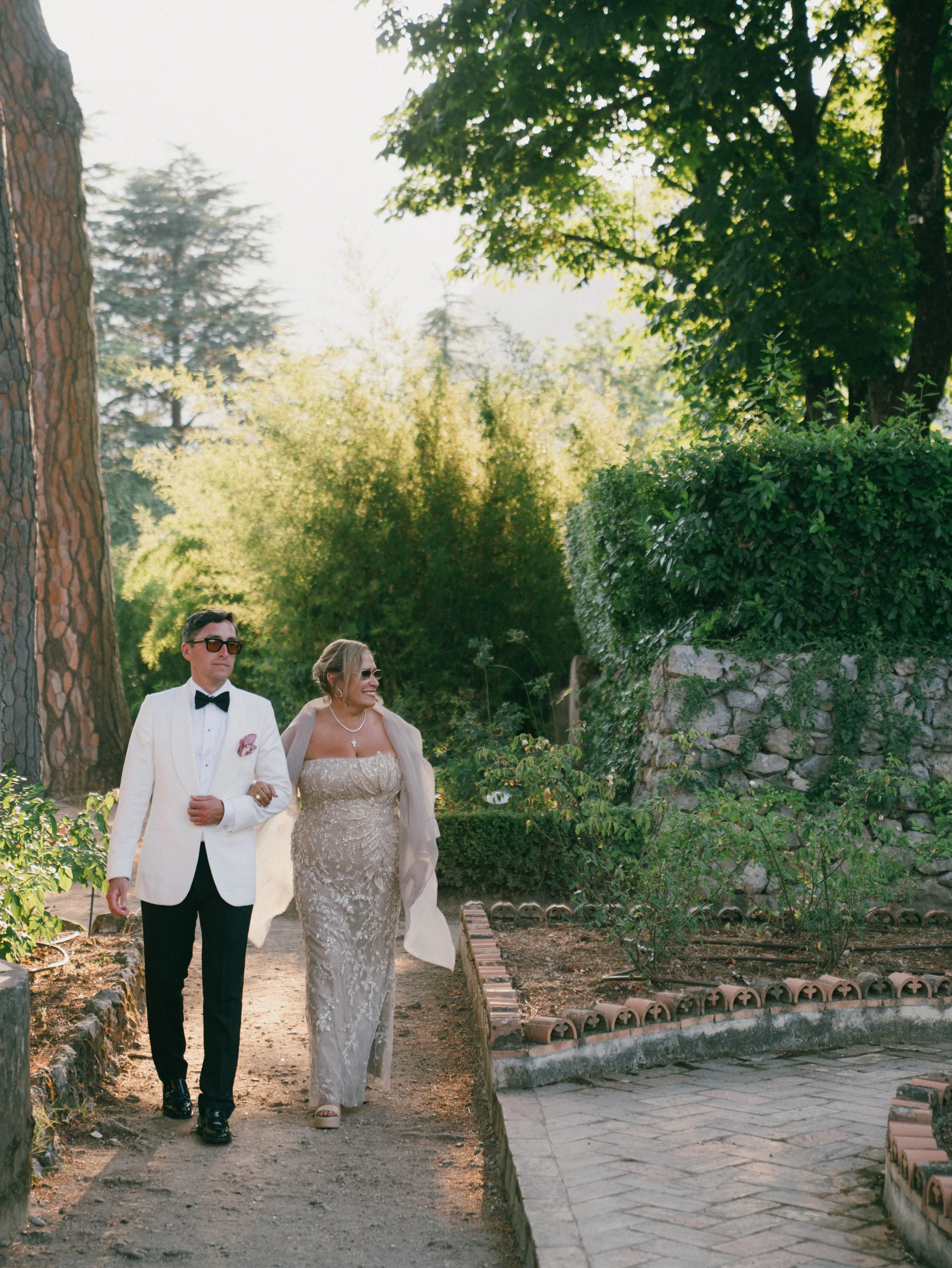A man in a tuxedo and a woman in a wedding dress walking outdoors in a garden with tall trees and greenery, smiling and holding hands.