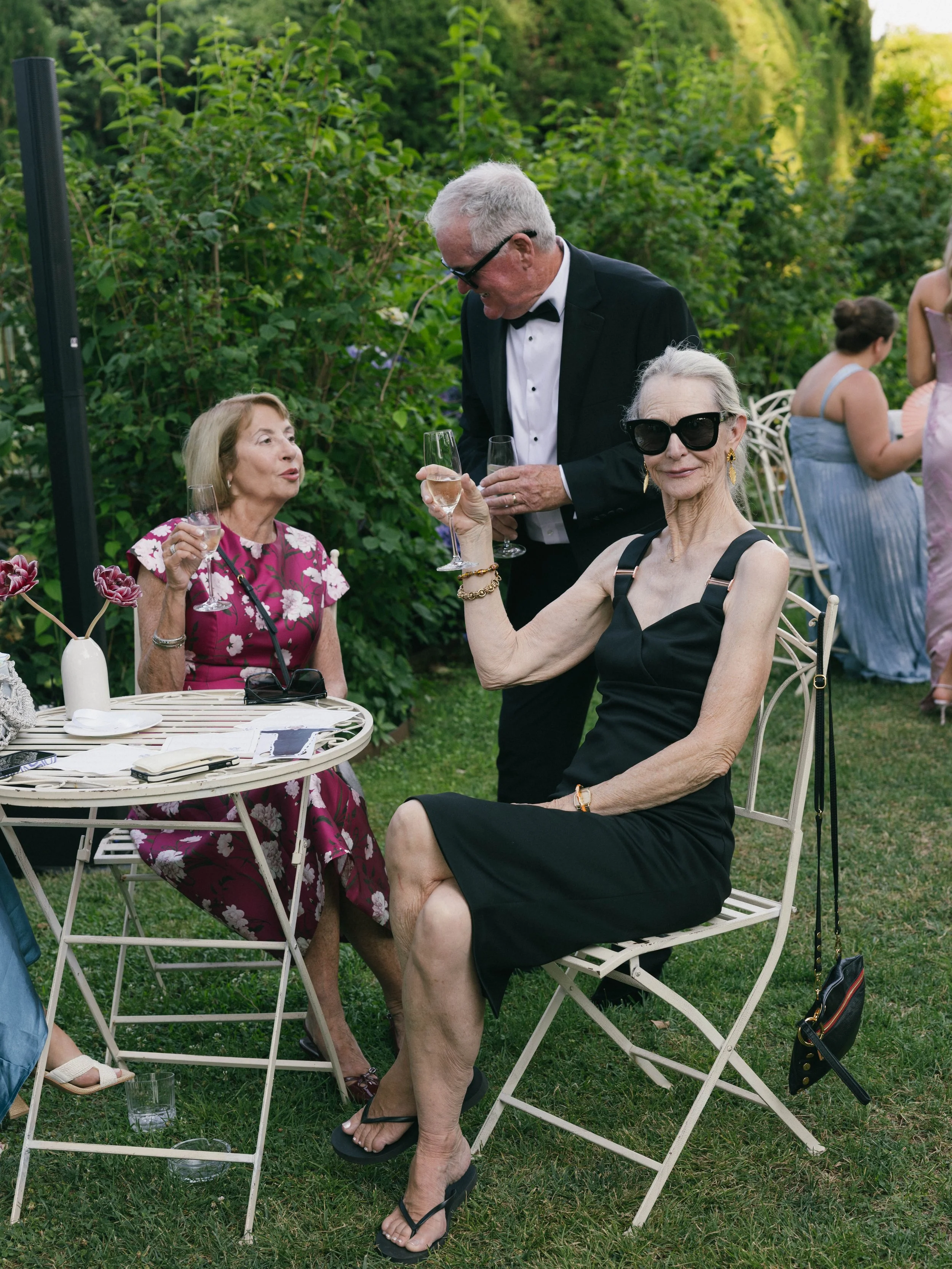 Older women and a man at an outdoor garden party, women sitting on chairs with glasses of champagne, man in tuxedo serving drinks, lush greenery background.