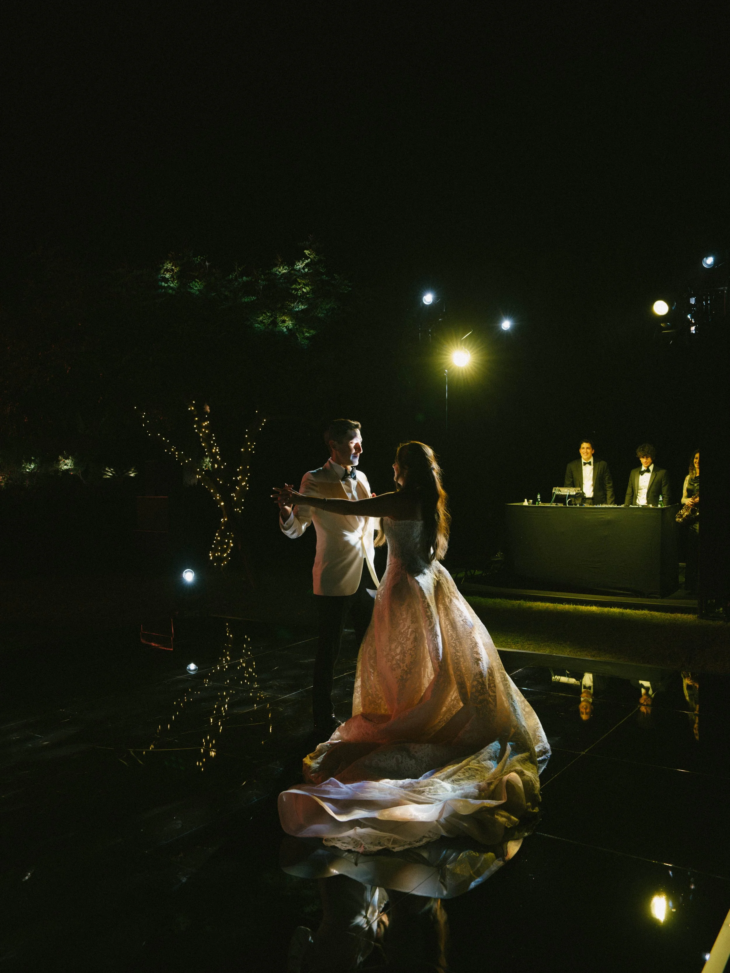 A couple is dancing at a nighttime outdoor wedding reception, illuminated by string lights and a few bright lights, with other guests and a DJ in the background.