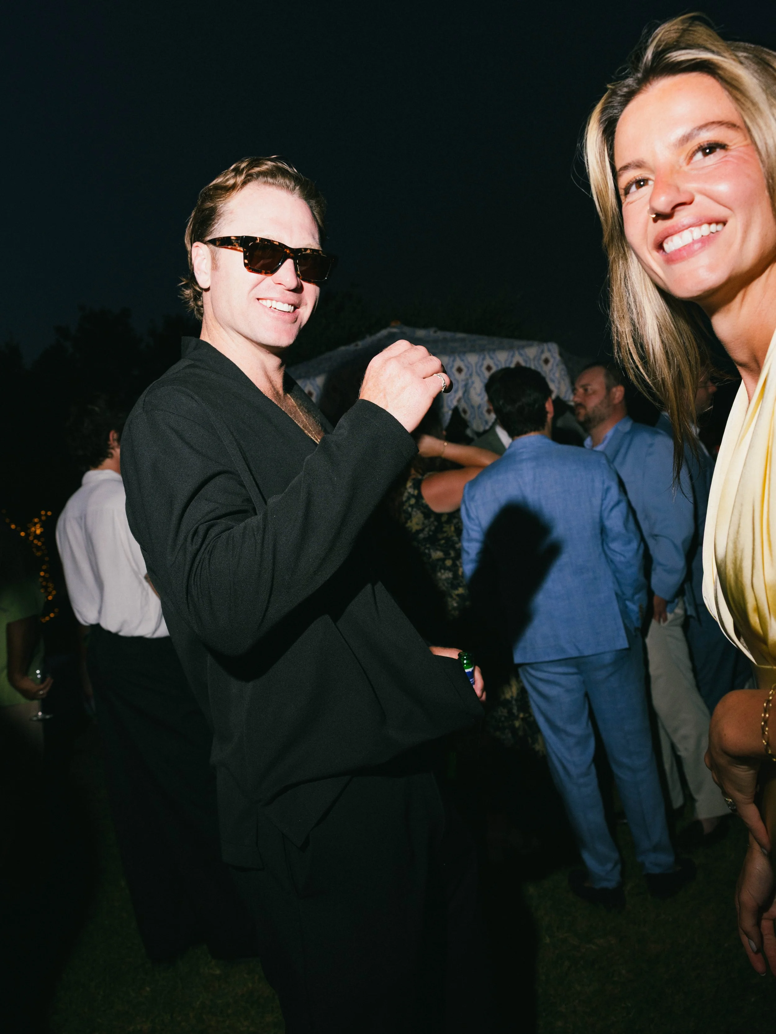 A man wearing sunglasses and a black blazer smiling at a party or social gathering at night, with other people in the background.