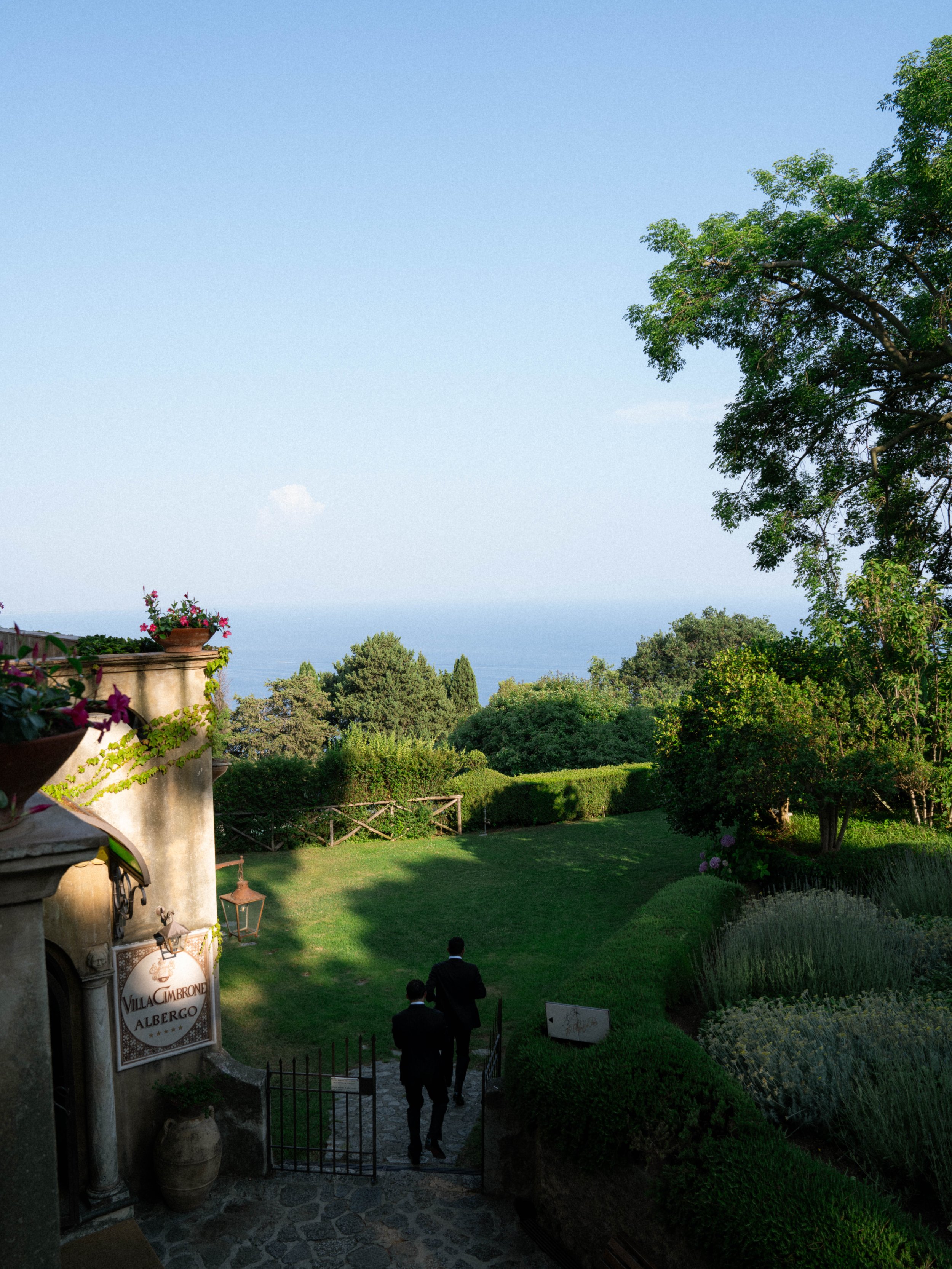 A view of a lush green garden with trees, shrubs, and flowering plants. Two men in suits are walking down a path towards an open gate, with a sign that reads 'Villa Cimbrone Albergo'. There is a large tree on the right and a blue sky overhead.