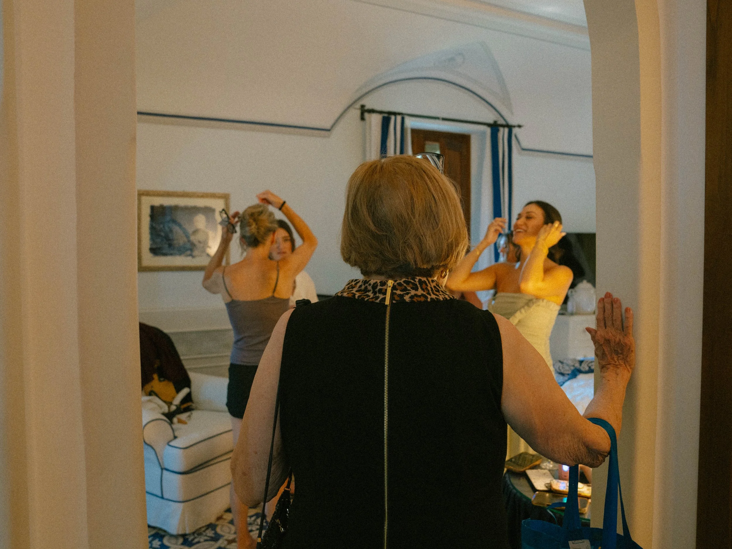 A woman with short red hair seen from behind adjusting her hair in front of a mirror, with two other women smiling and talking in the background.