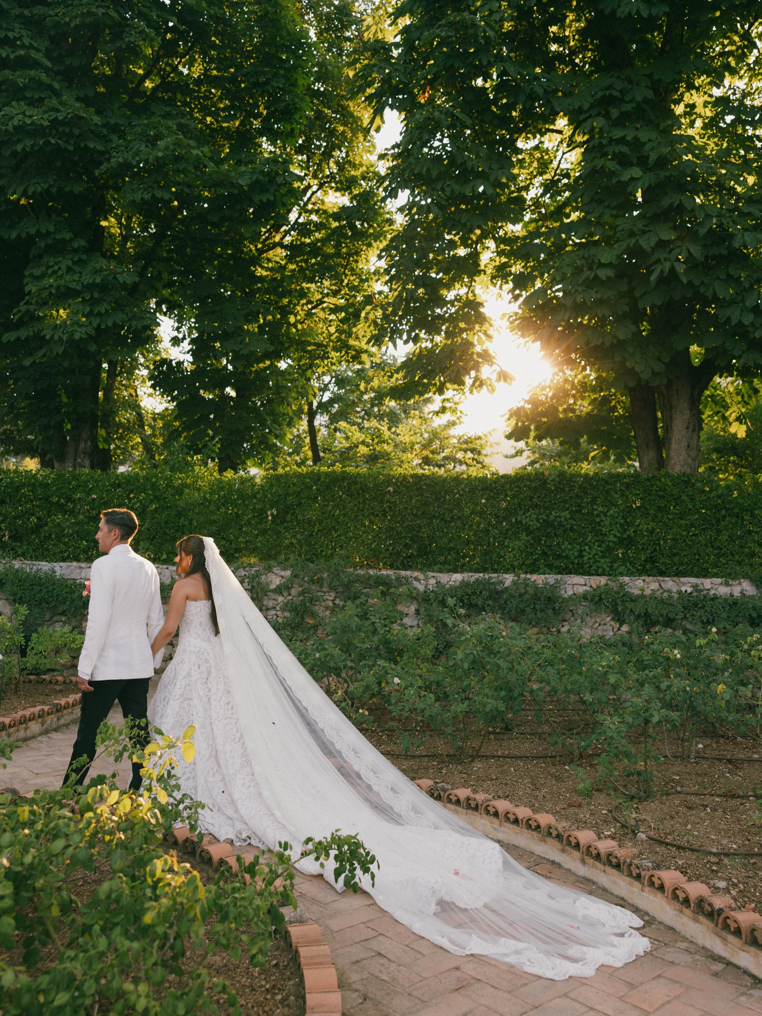 A bride and groom are walking outside during sunset, surrounded by trees and greenery, with the bride wearing a long wedding gown and veil, and the groom in a white jacket. Villa Cimbrone wedding Amalfi Coast