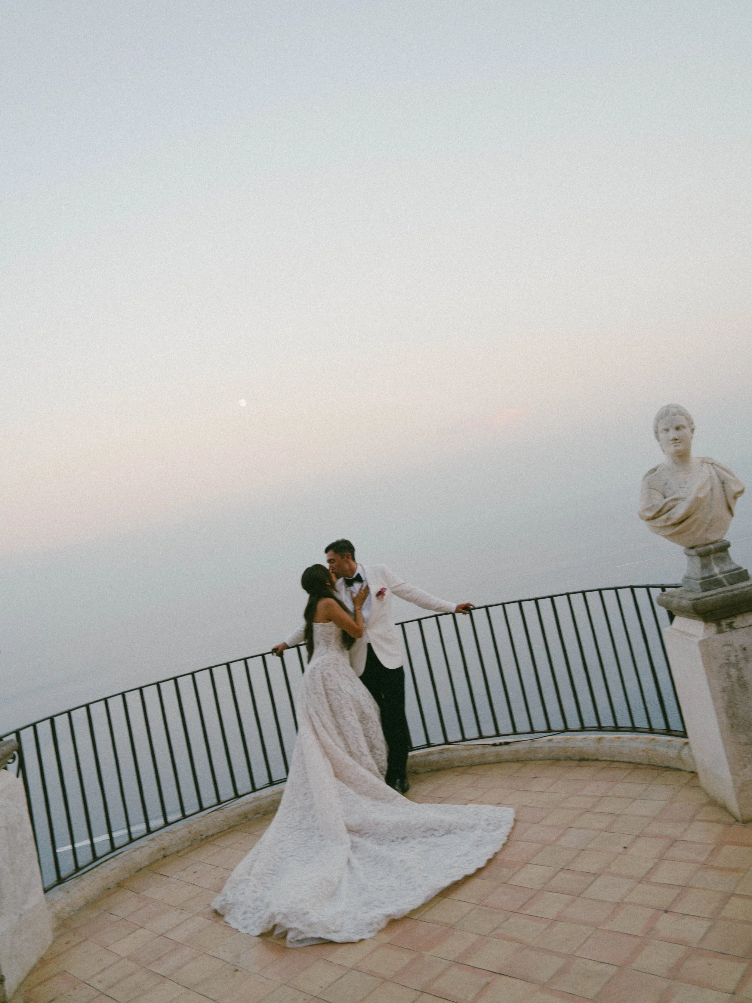 A bride and groom dancing outdoors on a terrace with a decorative railing, a large bust sculpture on a pedestal, and a clear sky with a hint of the moon in the background.