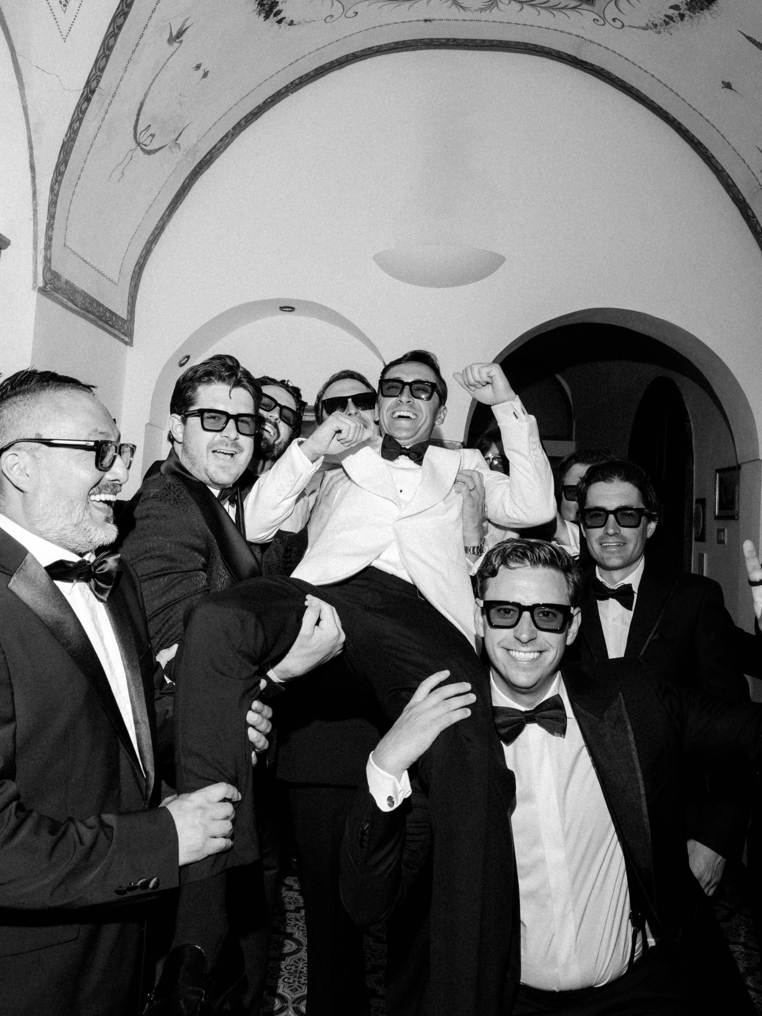 A group of men dressed in tuxedos and bow ties, some wearing glasses, celebrating and smiling at a formal event. They are posing together, with one man in the center raising his fist in victory or celebration. The setting appears to be an elegant ven