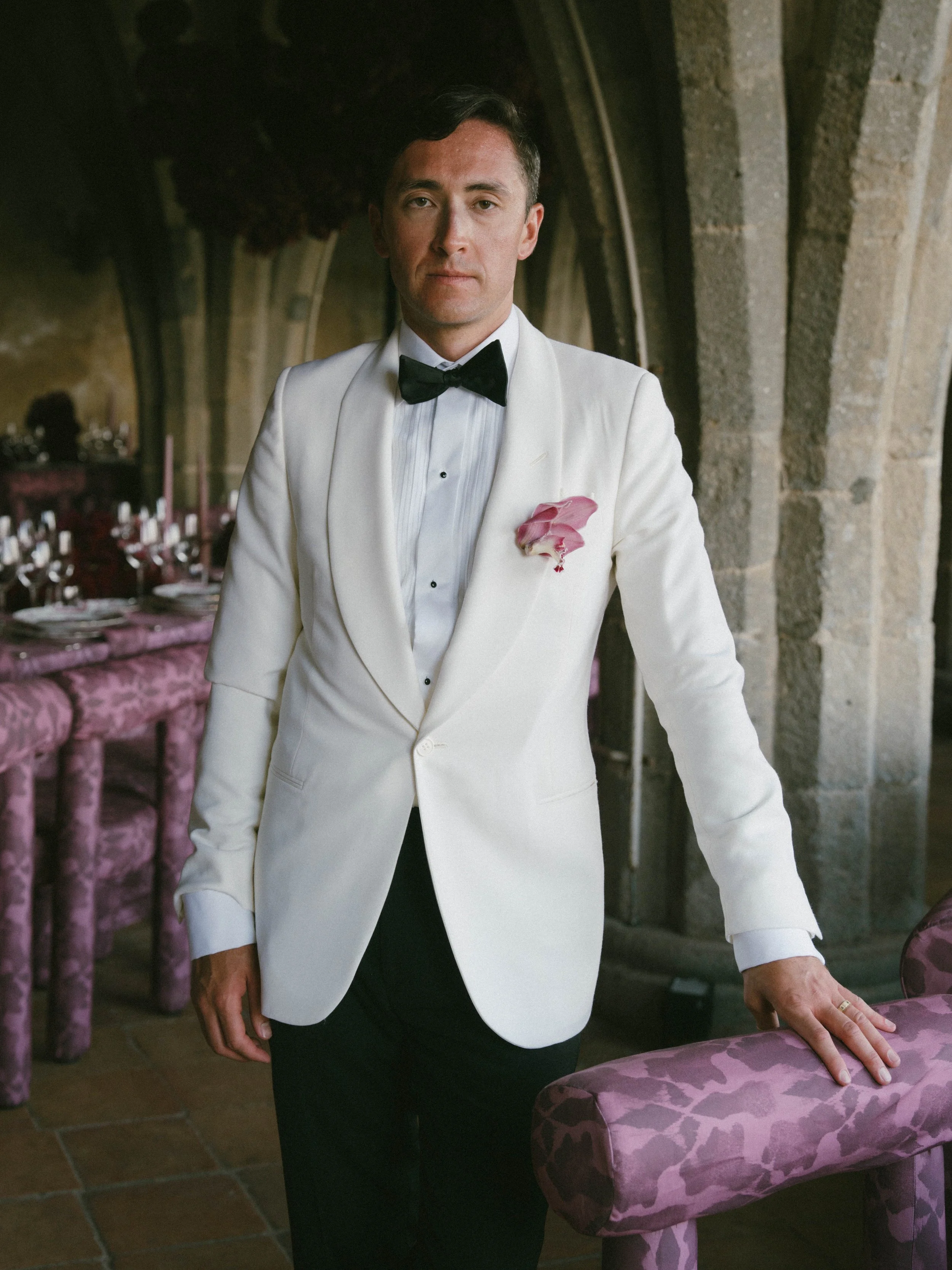 A man in a white tuxedo with a black bow tie and a pink flower on his lapel stands next to a purple patterned ottoman in a dimly lit setting resembling an elegant dining or banquet hall with stone arches.