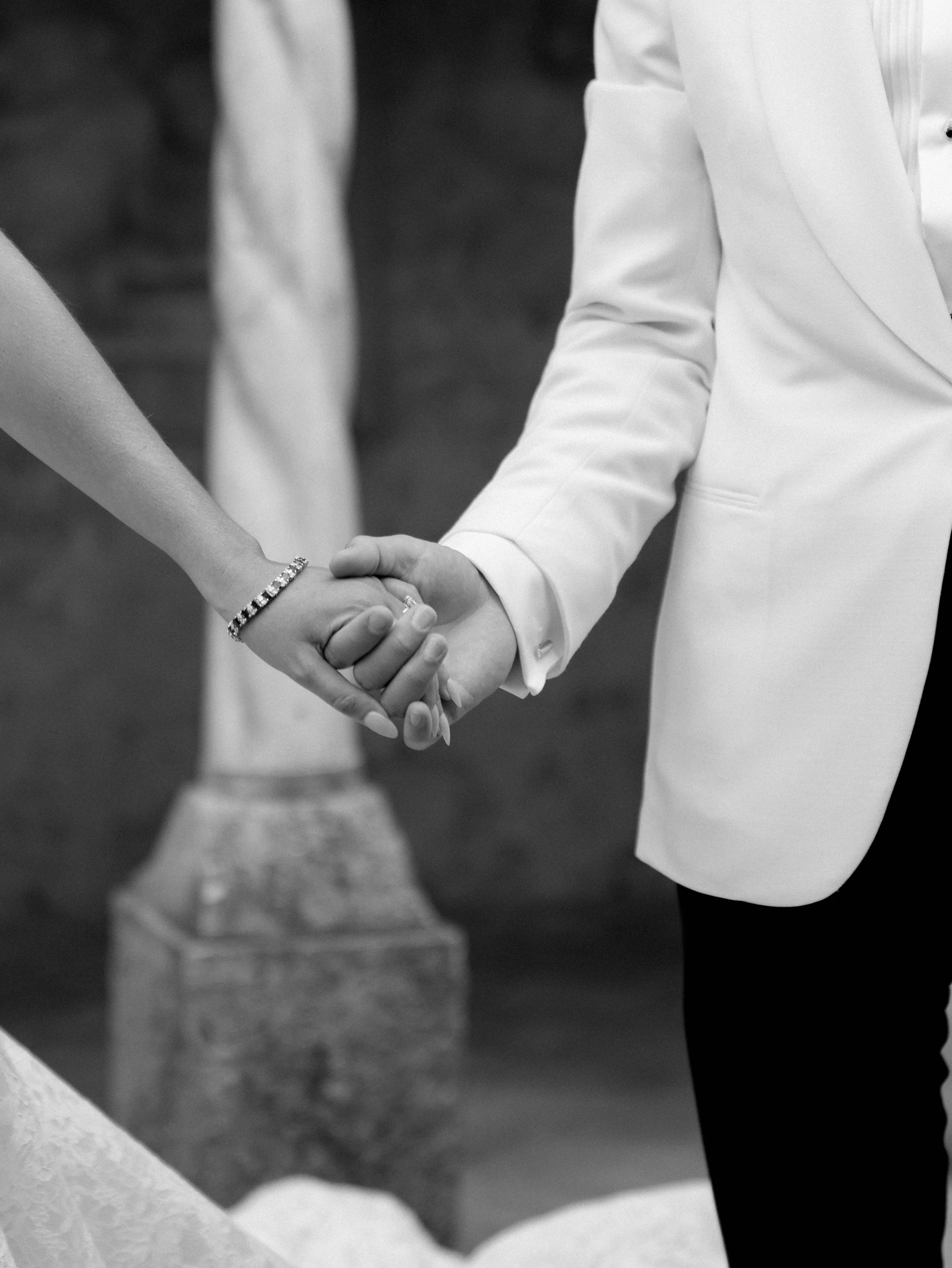 Close-up of a couple holding hands during a wedding ceremony, with bride wearing a bracelet and groom dressed in a suit, black and white photo.