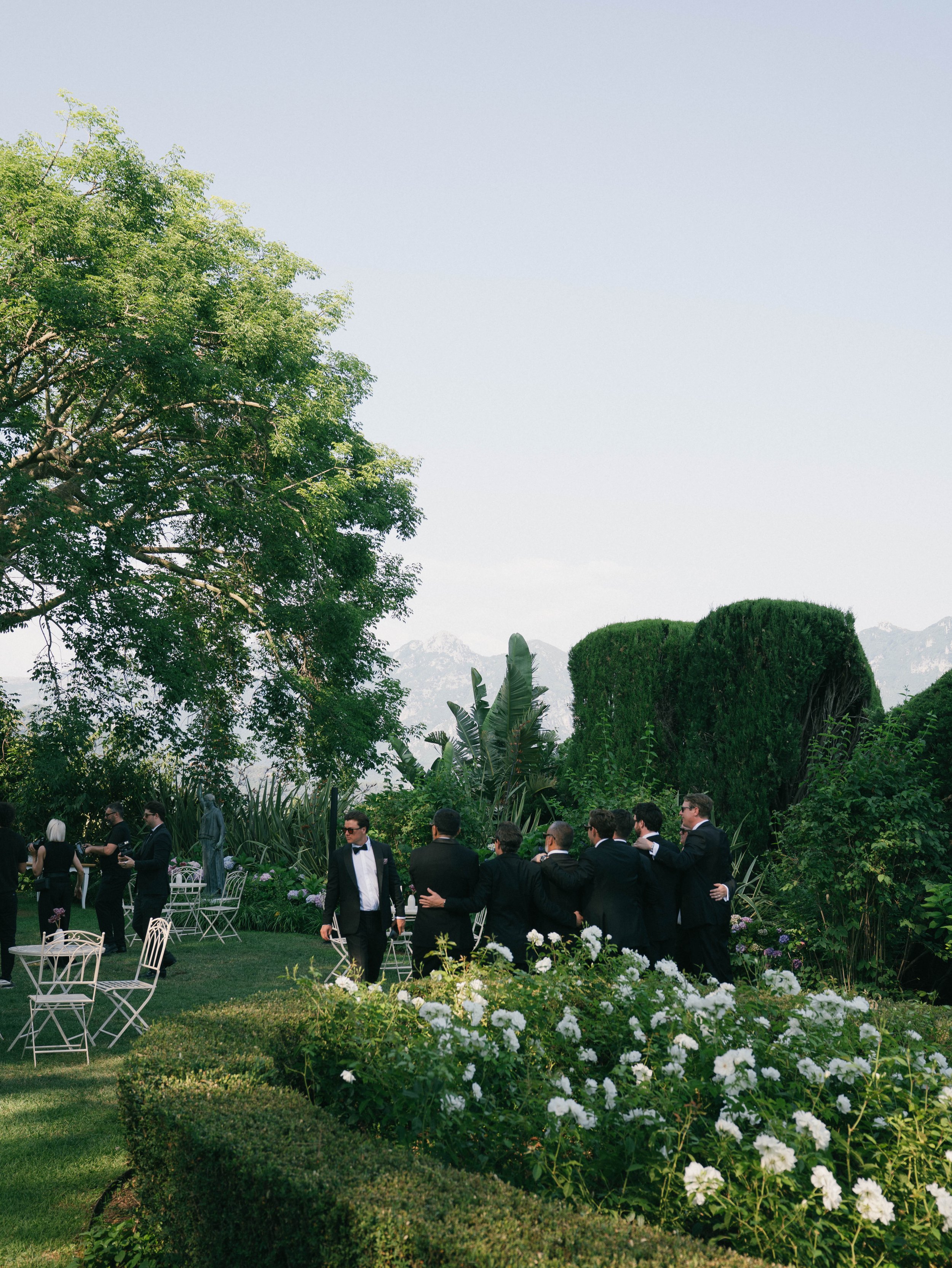 Group of men in formal attire at an outdoor garden wedding reception, with white chairs and lush greenery.