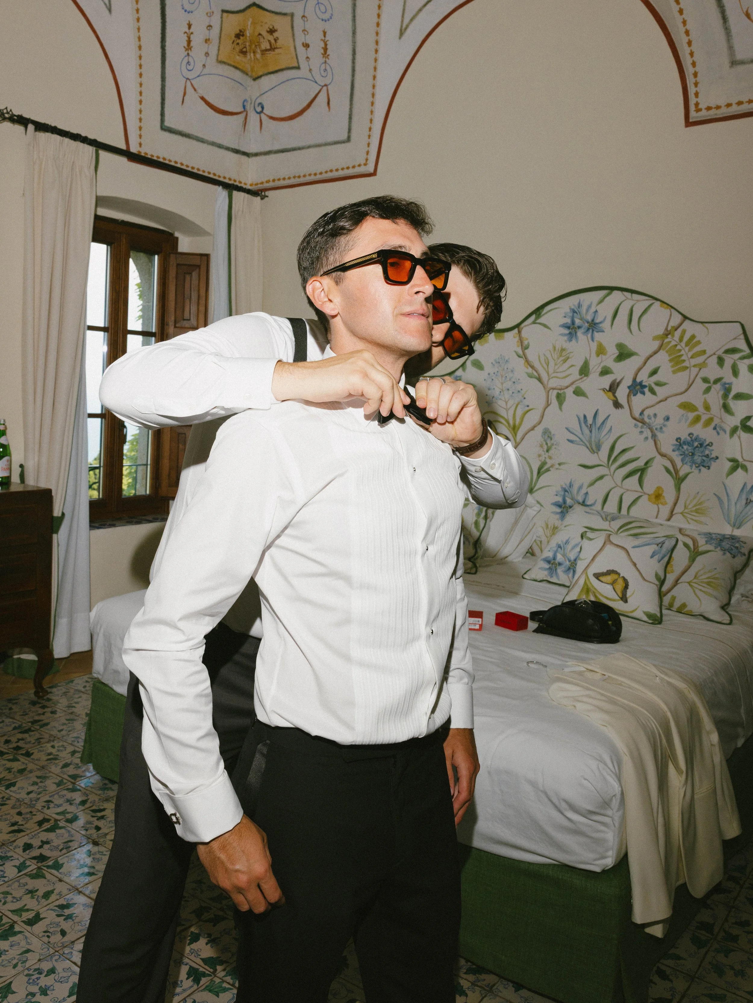 Two men dressed in formal attire wearing large, tinted sunglasses, standing in a bedroom with floral patterned walls and ceiling paintings, with one man in the foreground adjusting his collar.