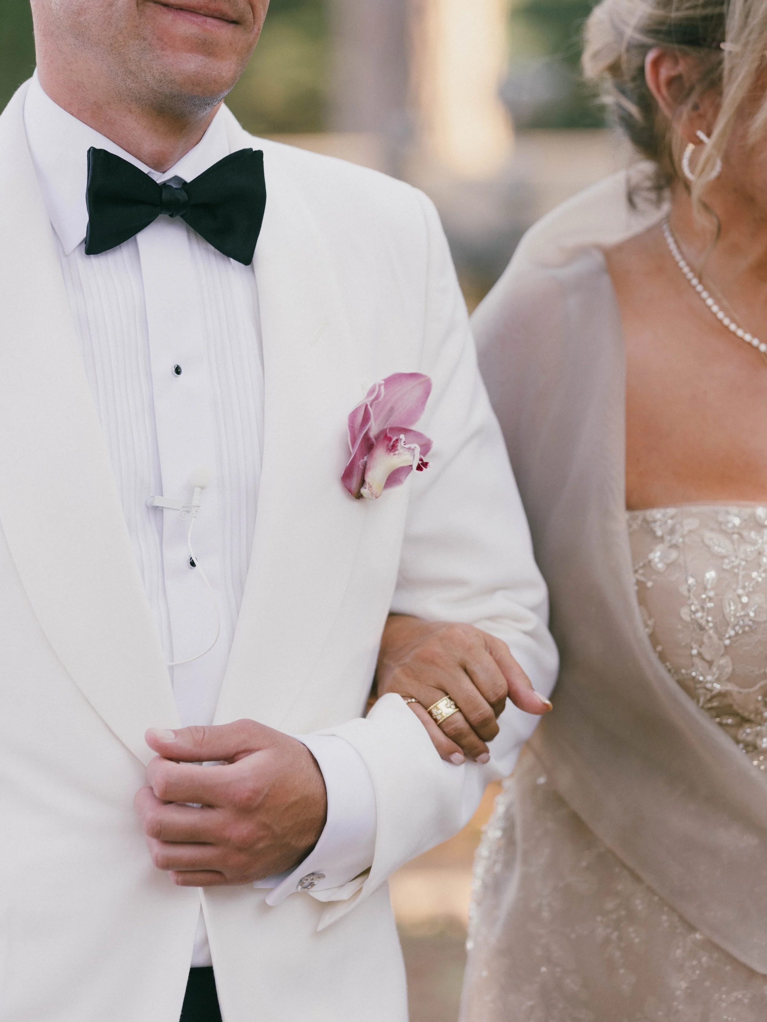 Close-up of a groom in a white tuxedo with a black bow tie and a pink orchid boutonniere, standing next to a bride in a wedding dress with lace and bead details, both with arms crossed, at an outdoor wedding.