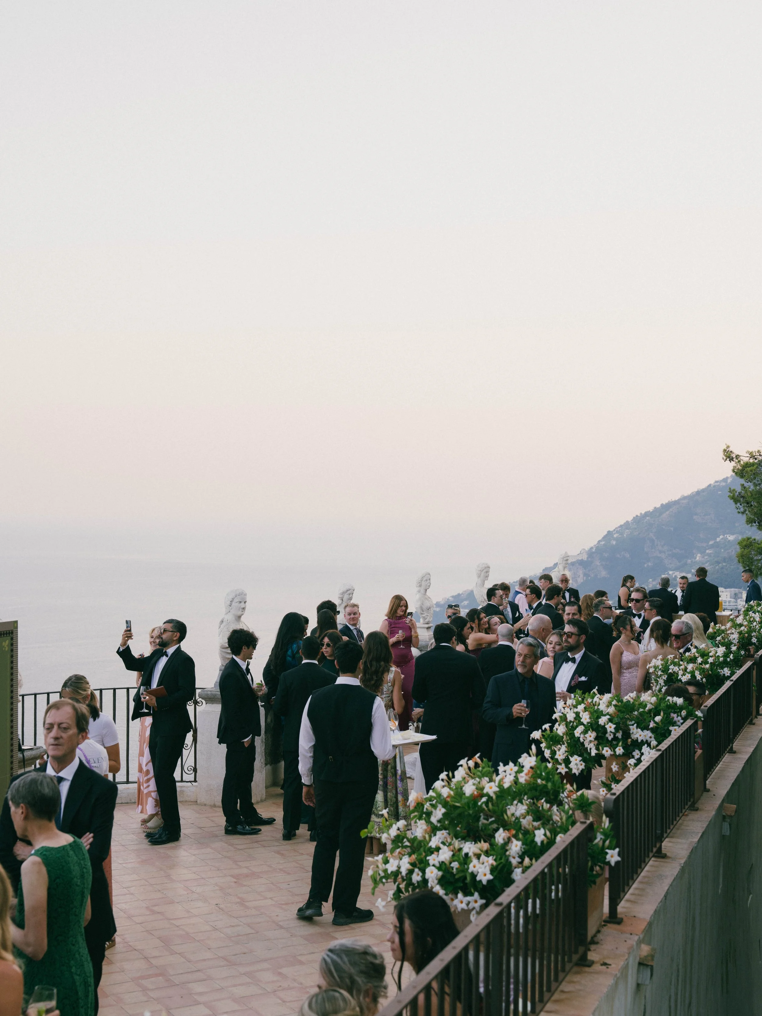 People dressed formally attending a social event on a terrace overlooking a body of water, with a mountain in the background and flowers in the foreground. Villa Cimbrone wedding Amalfi Coast Ravello Infinity Terrace