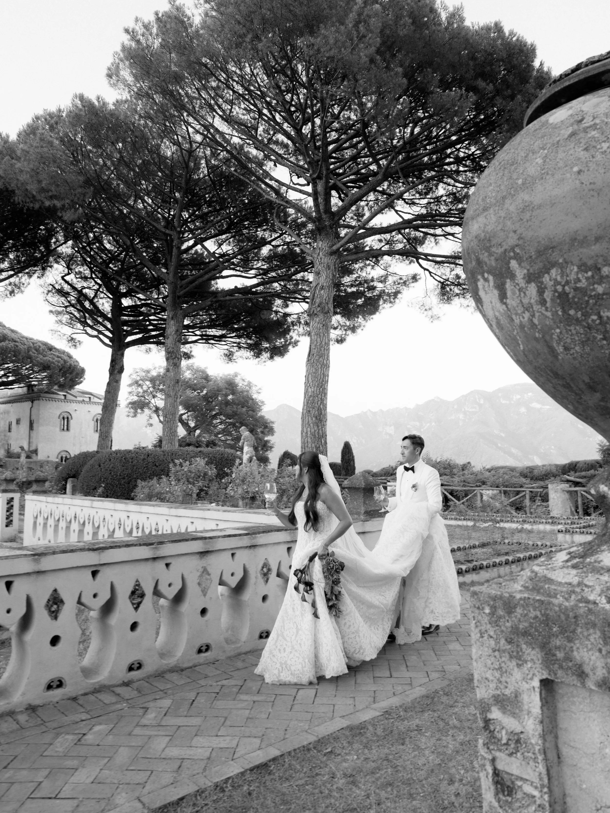 A black and white photo of a bride and groom in wedding attire walking outdoors, with large trees in the background, a decorative cast-iron fence, and distant mountains. Villa Cimbrone wedding Amalfi Coast Ravello