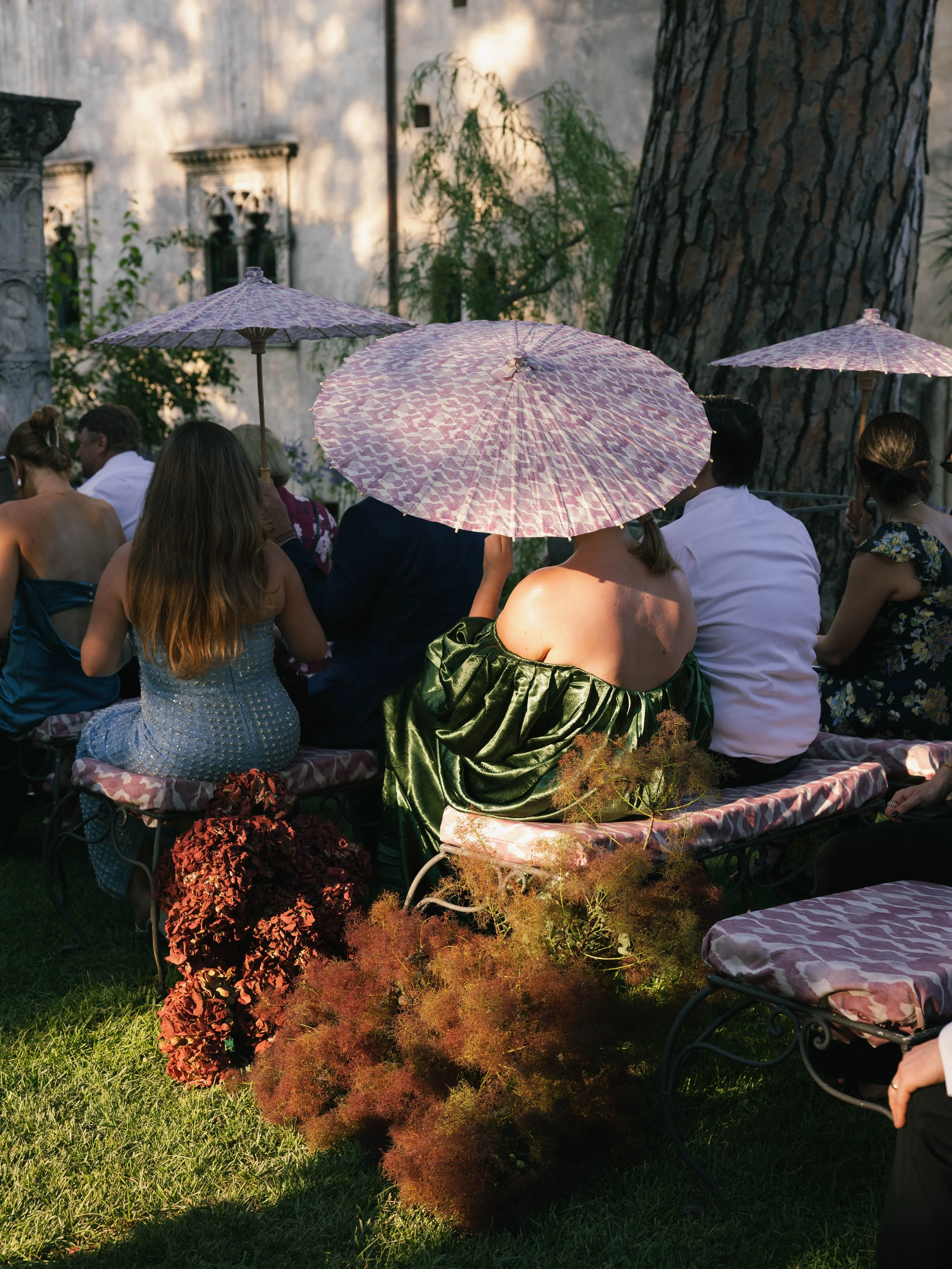 People sitting outdoors with pink parasols and floral patterned chairs, with greenery and a large tree in the background.