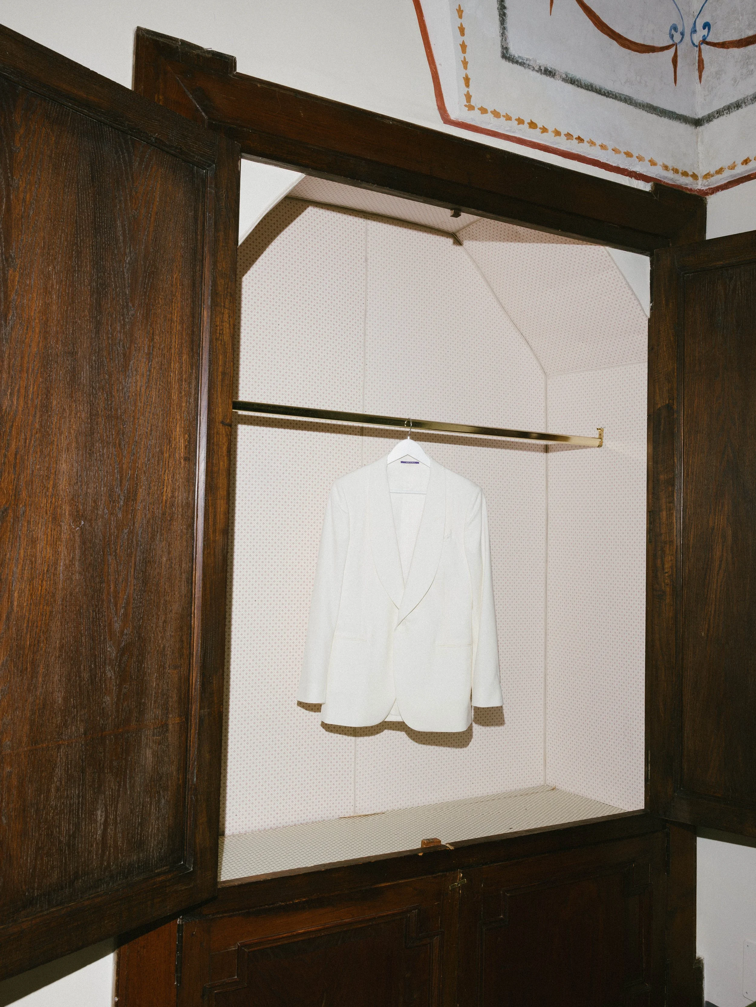 A white blazer hanging on a clothes rack inside a wooden display cabinet with open doors, against a beige and patterned wall.