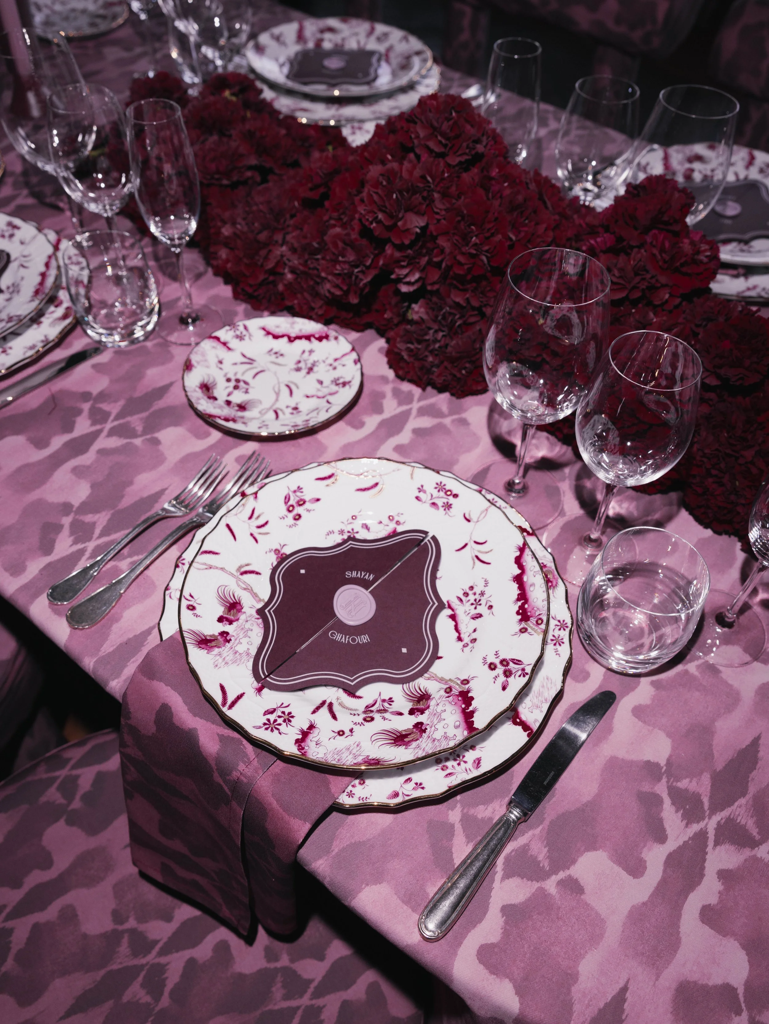 Elegant table setting with floral-patterned plates, wine glasses, silverware, and a pink floral tablecloth, featuring a centerpiece of deep red flowers.Villa Cimbrone wedding Amalfi Coast