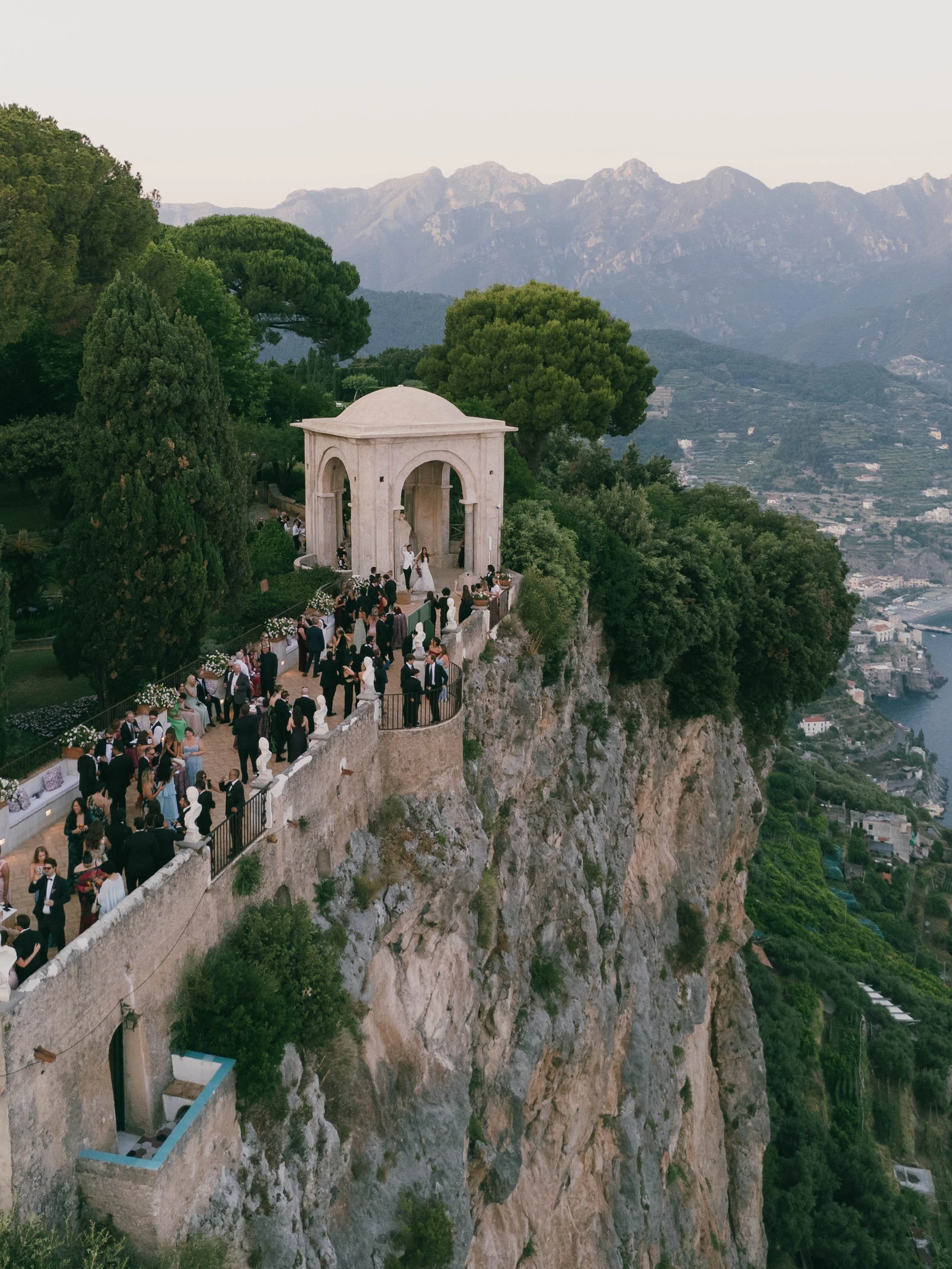 A large group of people gathered at a wedding ceremony on a platform built into a cliffside with a small white pavilion in the background. The scene overlooks a valley with mountains in the distance at the base of the mountains. Villa Cimbrone, Amalf