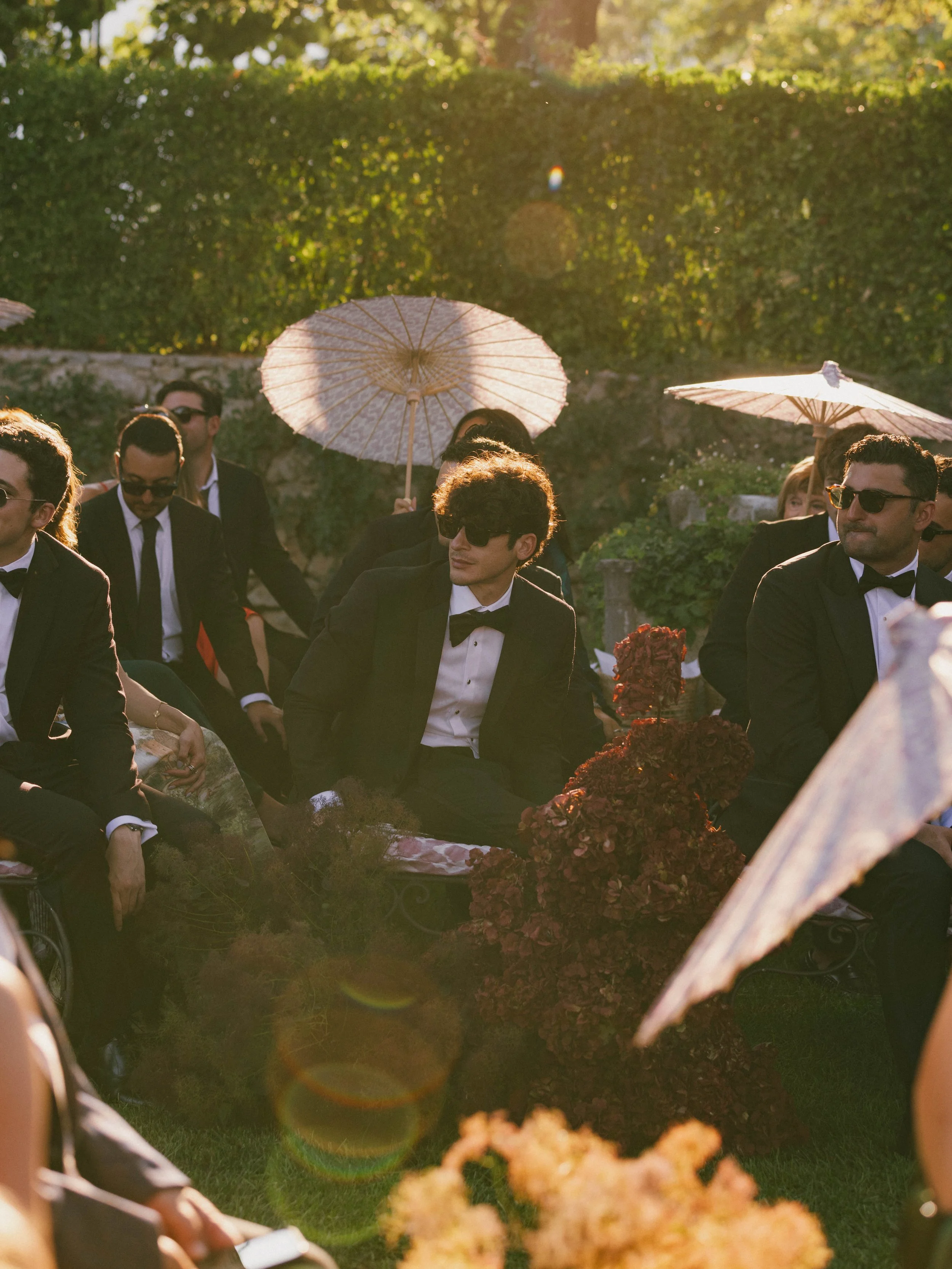 People dressed in formal attire, some holding parasols, seated outdoors in a garden with sunlight. Villa Cimbrone wedding Amalfi Coast