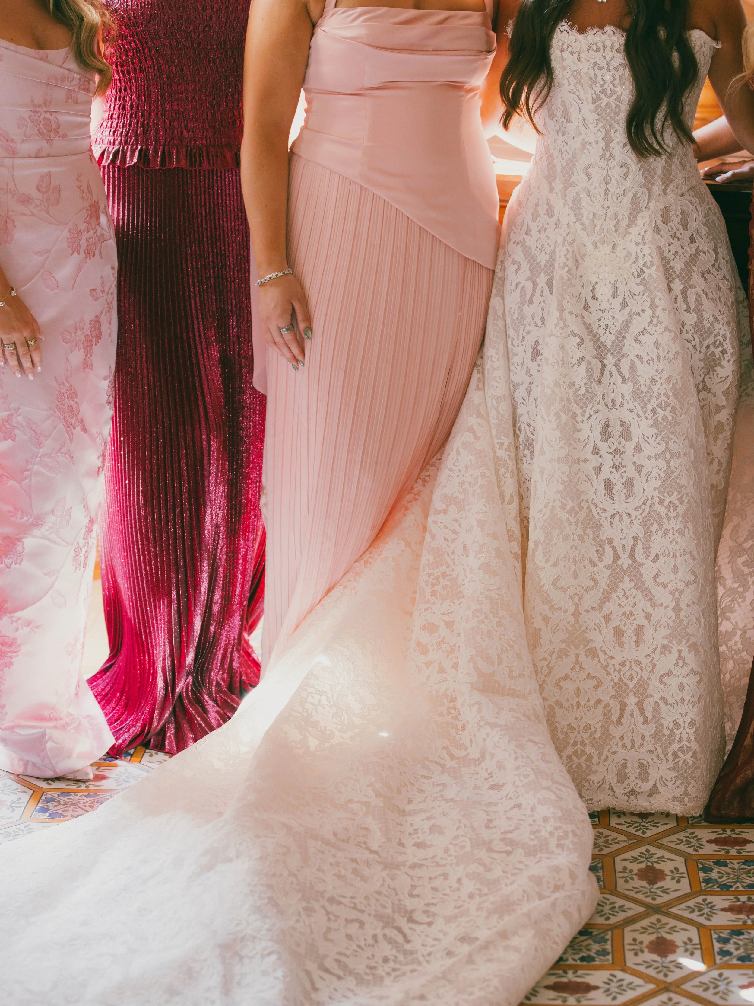 Close-up of women wearing elegant dresses, including pink, red, and white lace, at a formal event, with intricate patterned flooring visible at the bottom.