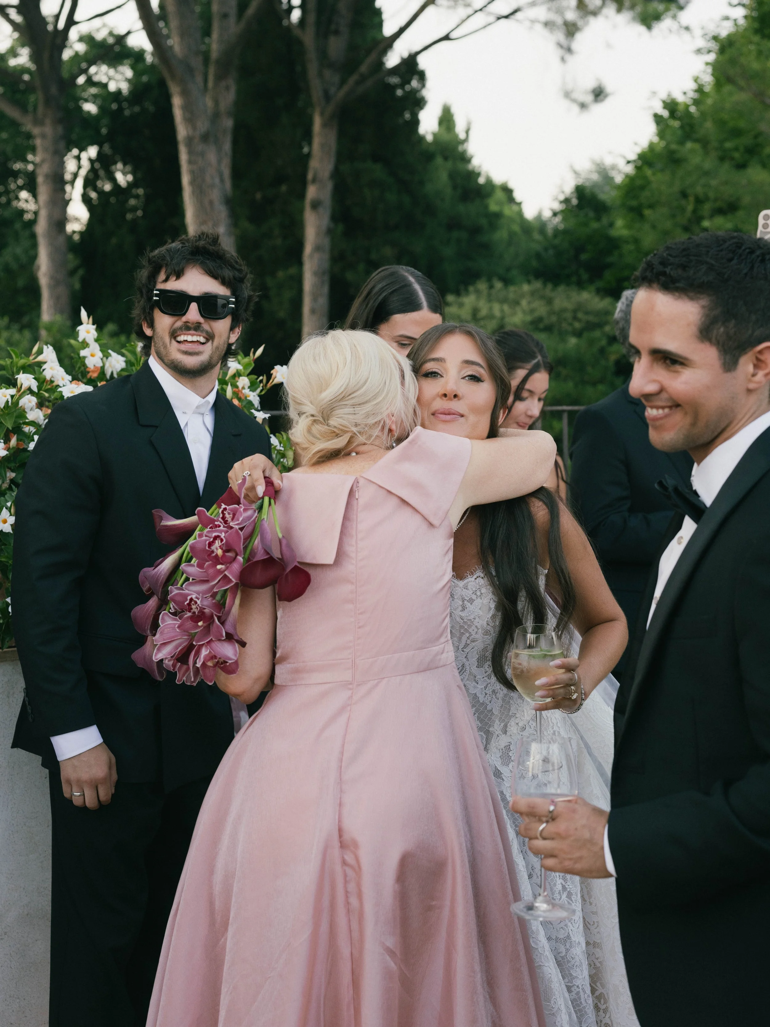 Group of people celebrating outdoors, embracing, smiling, holding glasses of wine, with trees and greenery in the background. Villa Cimbrone wedding Amalfi Coast