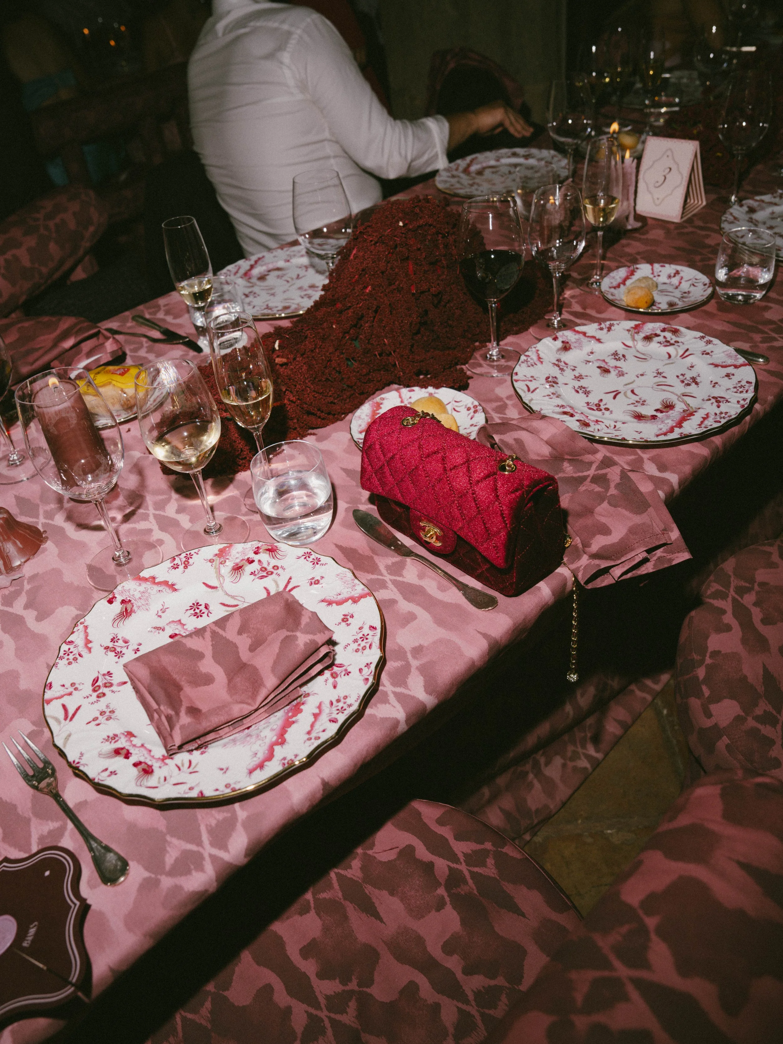 A dinner table set for a celebration with pink tablecloth, floral plates, champagne glasses, and a red quilted purse, with a person in a white shirt sitting in the background.