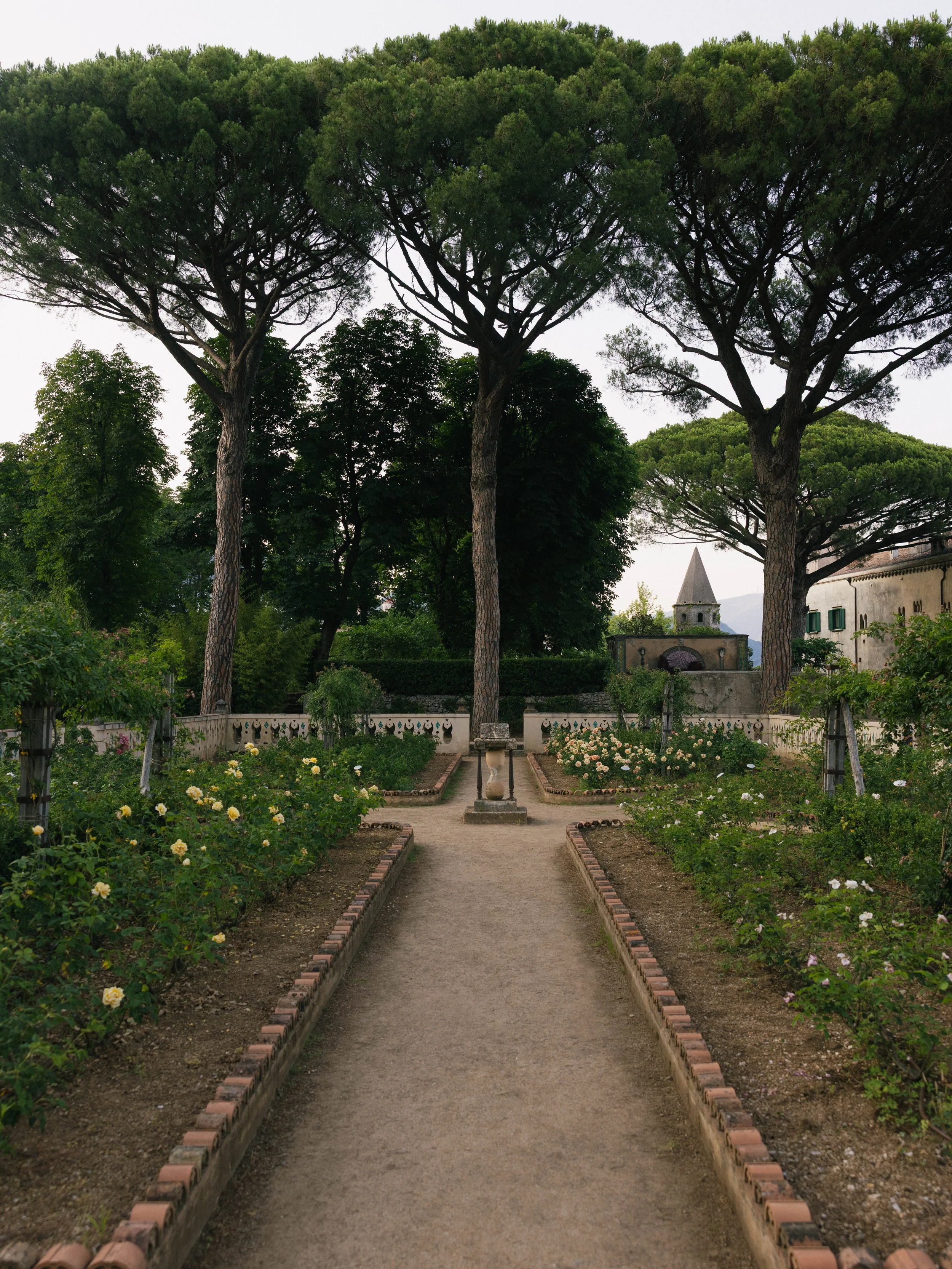 A garden path lined with flowers and small bricks leading to trees and a building with a pointed roof, under a cloudy sky. Villa Cimbrone wedding Amalfi Coast