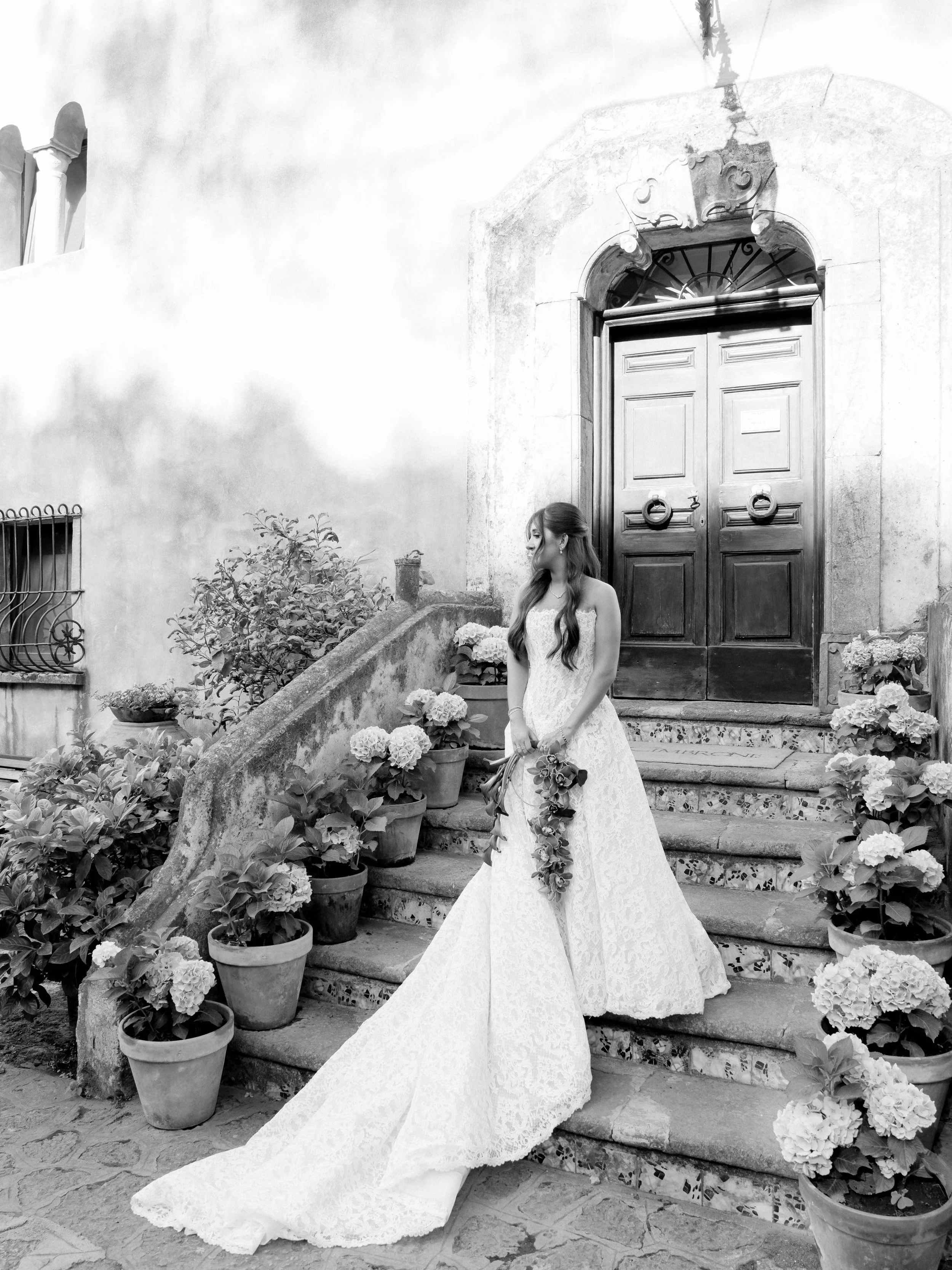 A woman in a wedding dress standing on stone steps outside a building with large wooden doors, surrounded by potted hydrangeas.