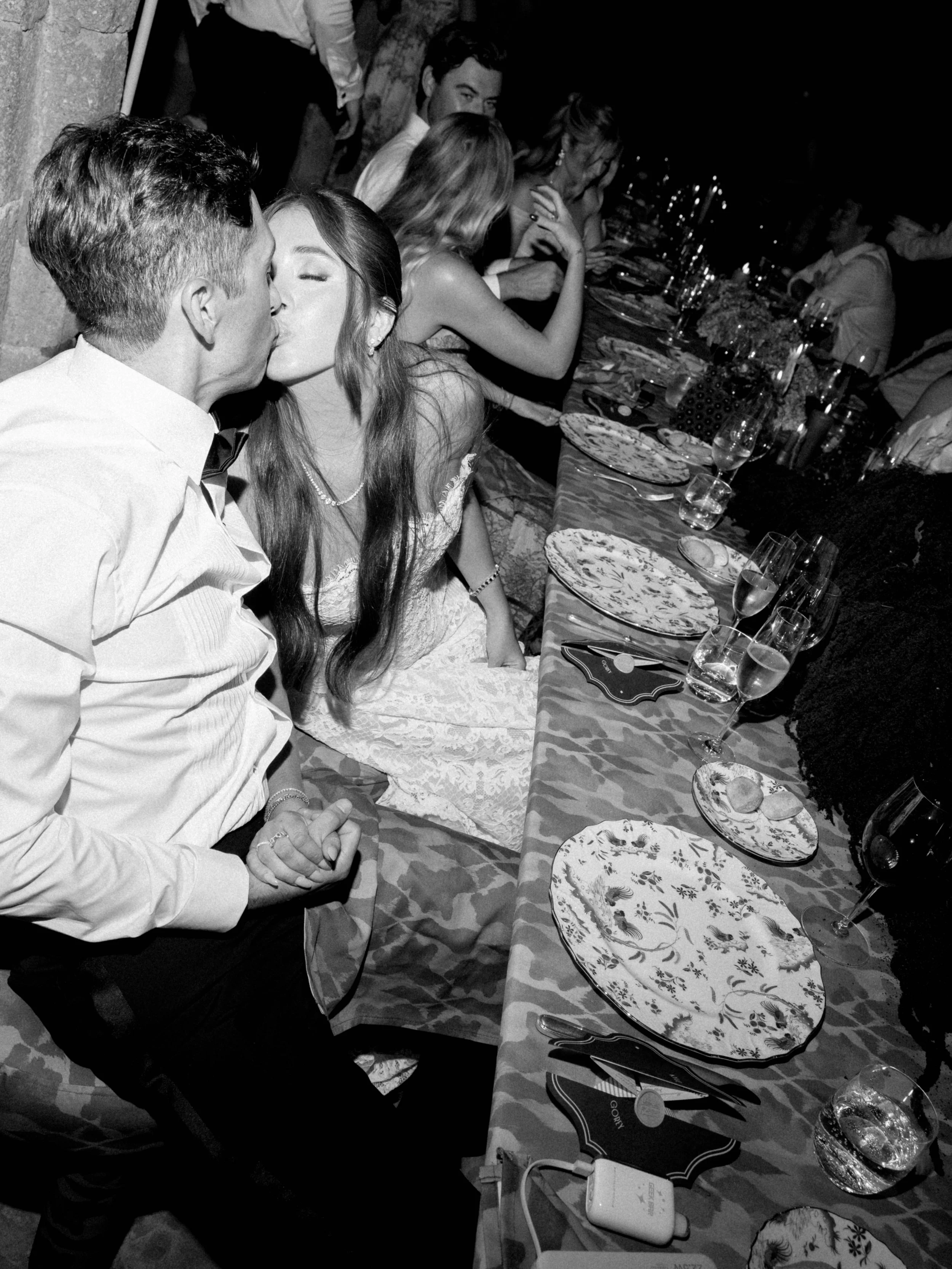 A black and white photo of a dinner party with several people seated along a long table, some drinking, talking, and one couple in the foreground sharing a kiss.