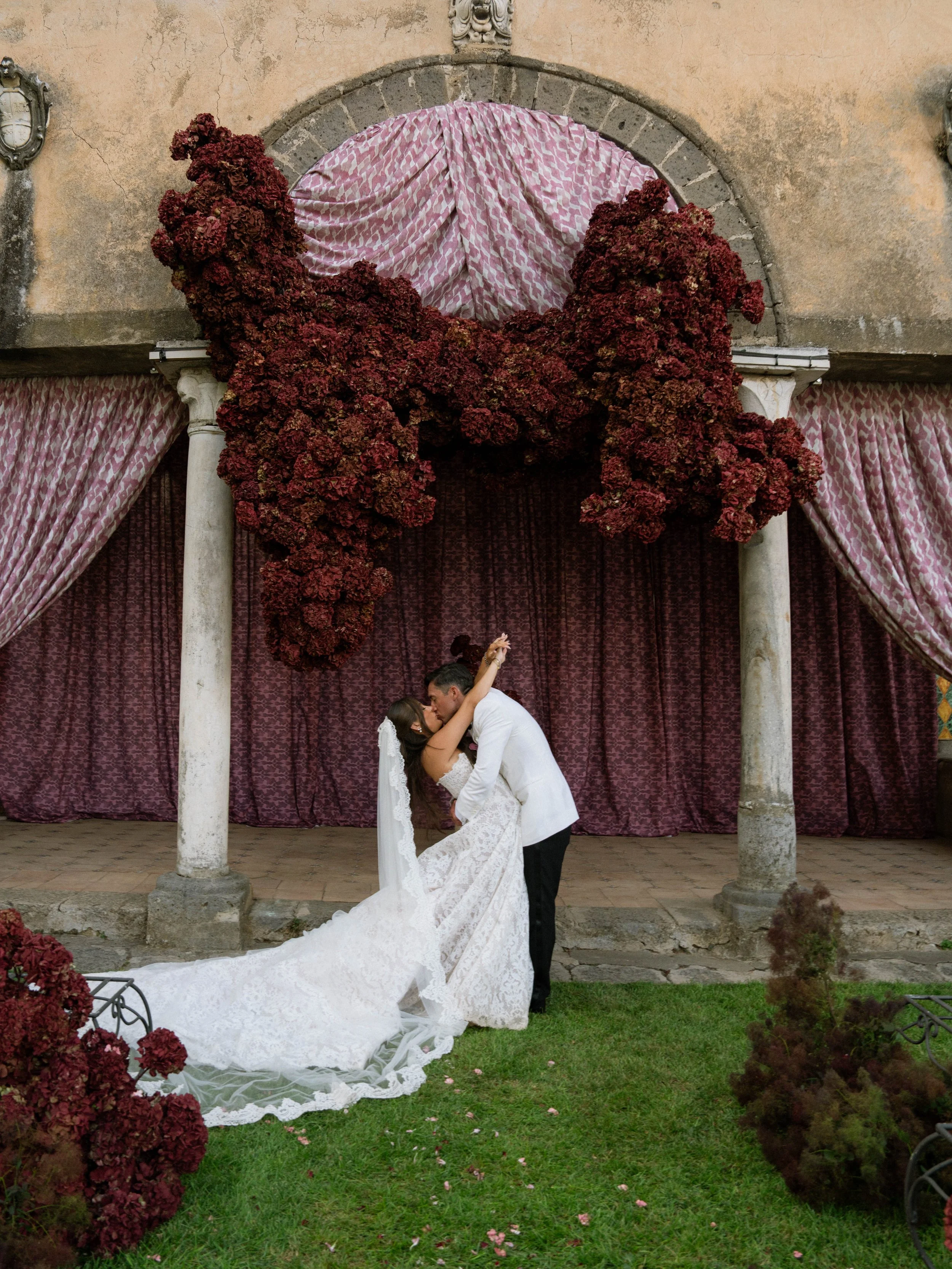 A bride and groom kiss under a floral arch with purple curtains and stone columns, surrounded by greenery and flowers. Villa Cimbrone wedding Amalfi Coast