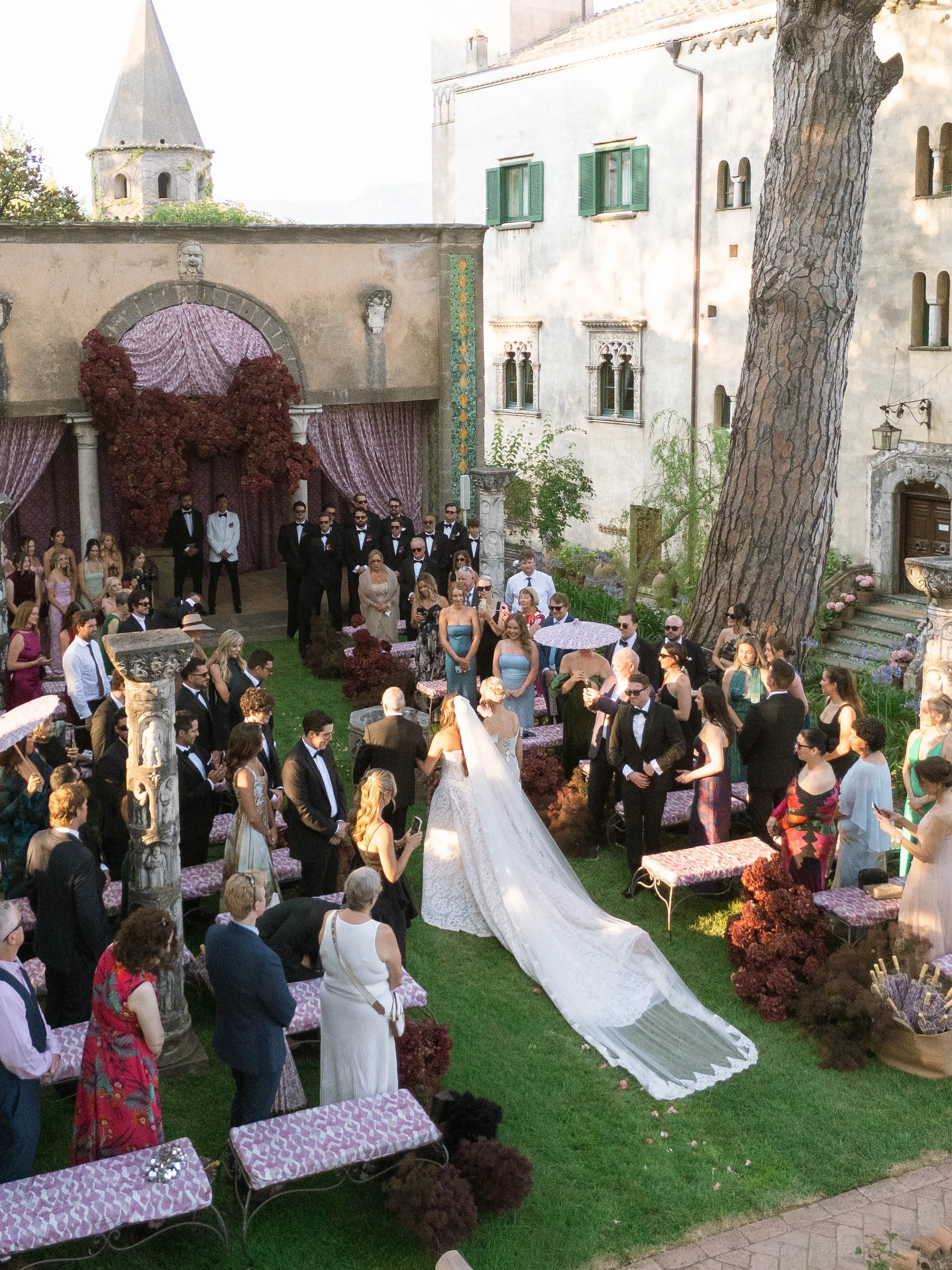 A wedding celebration in a garden with guests standing and sitting, a bride in a white gown, a groom, and a group of men in tuxedos. There are tables with purple patterned tablecloths, a large tree, and a stone building with green-shuttered windows i