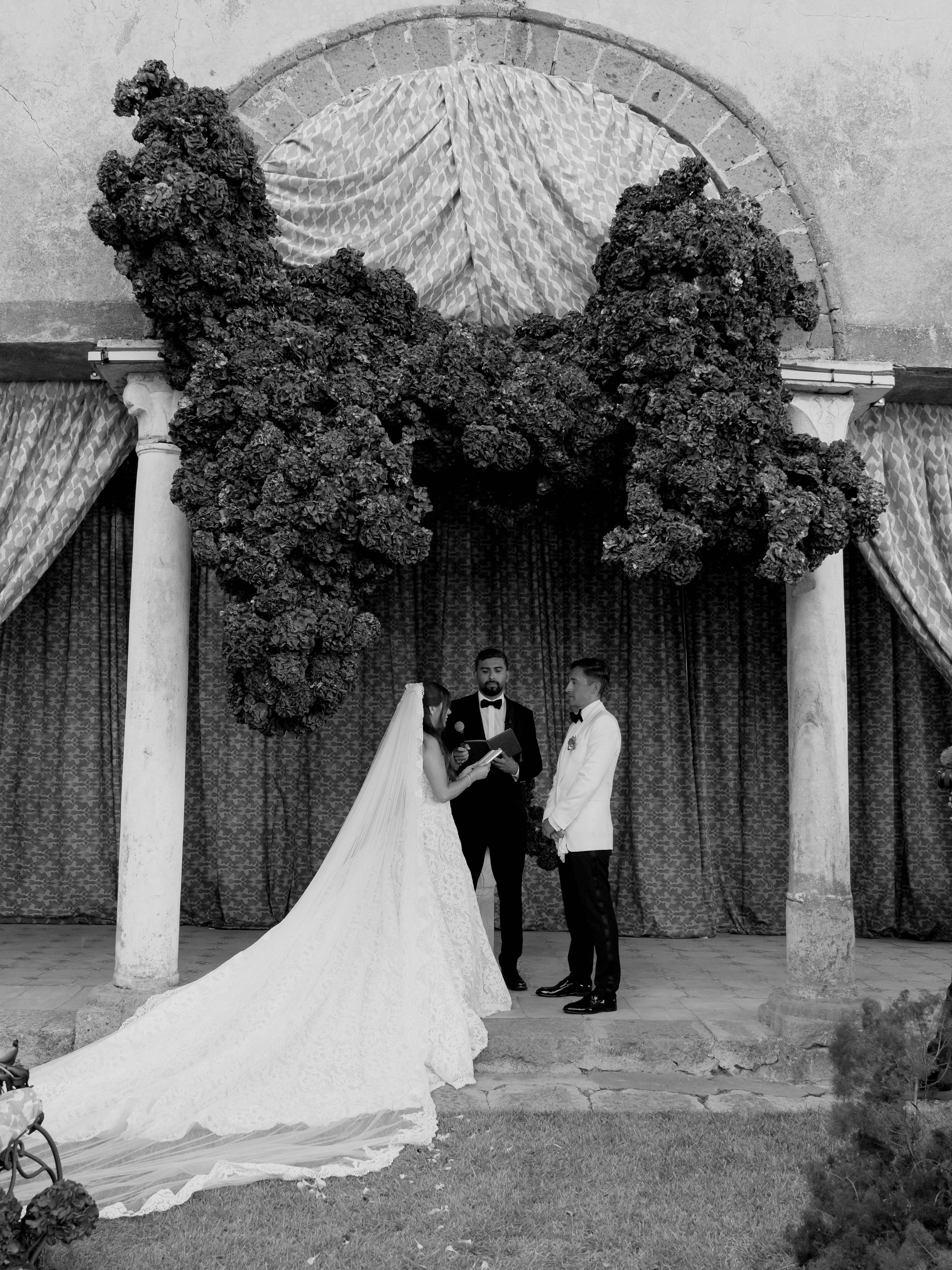 A wedding ceremony under an arch decorated with large floral arrangements. The bride and groom stand together with a person officiating the ceremony, possibly a priest or officiant. The scene is outdoors, with curtains or drapes hanging in the backgr
