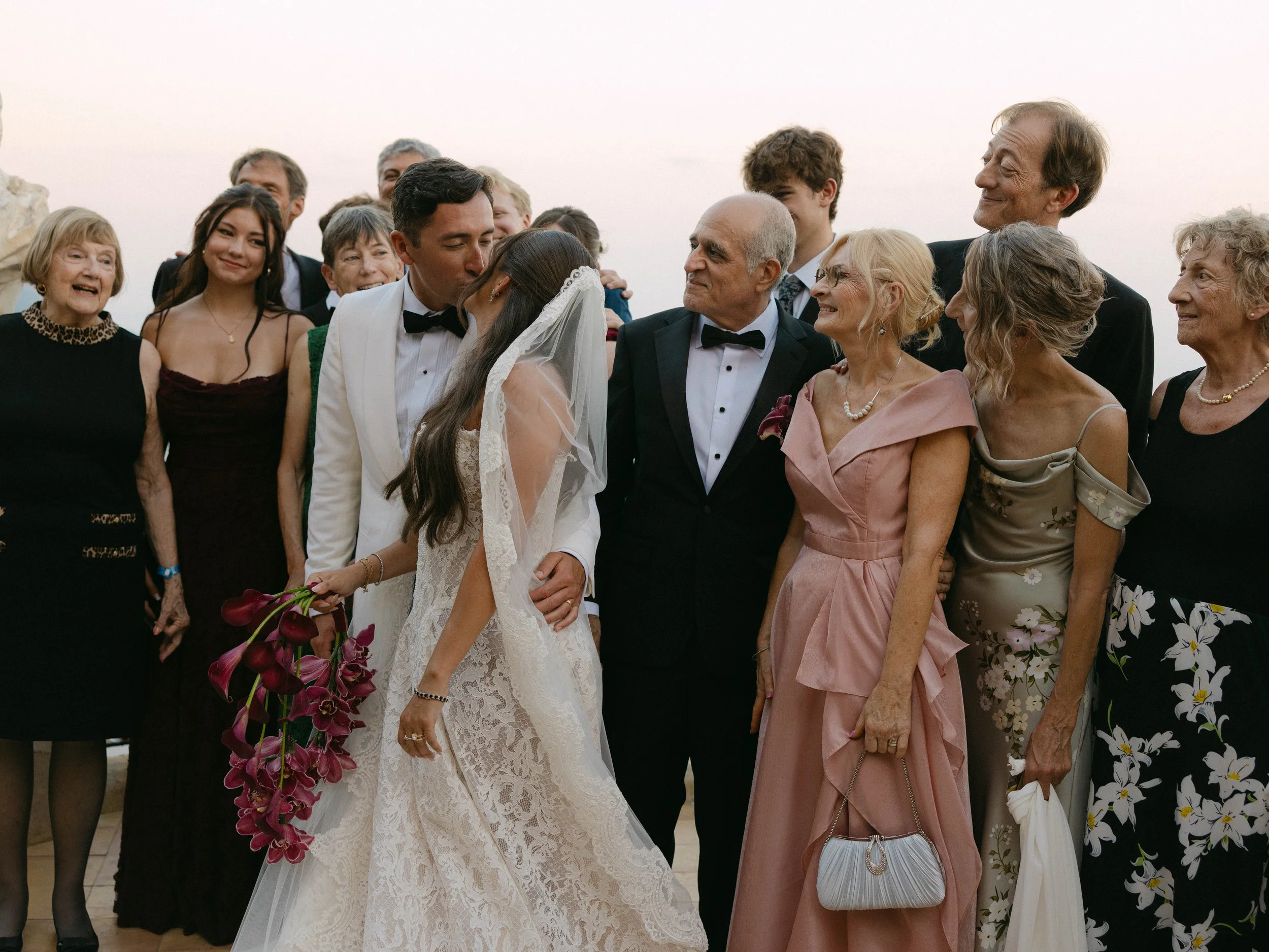 A group of diverse people gathered outdoors, with a bride and groom in the center, kissing during a wedding celebration surrounded by family and friends.