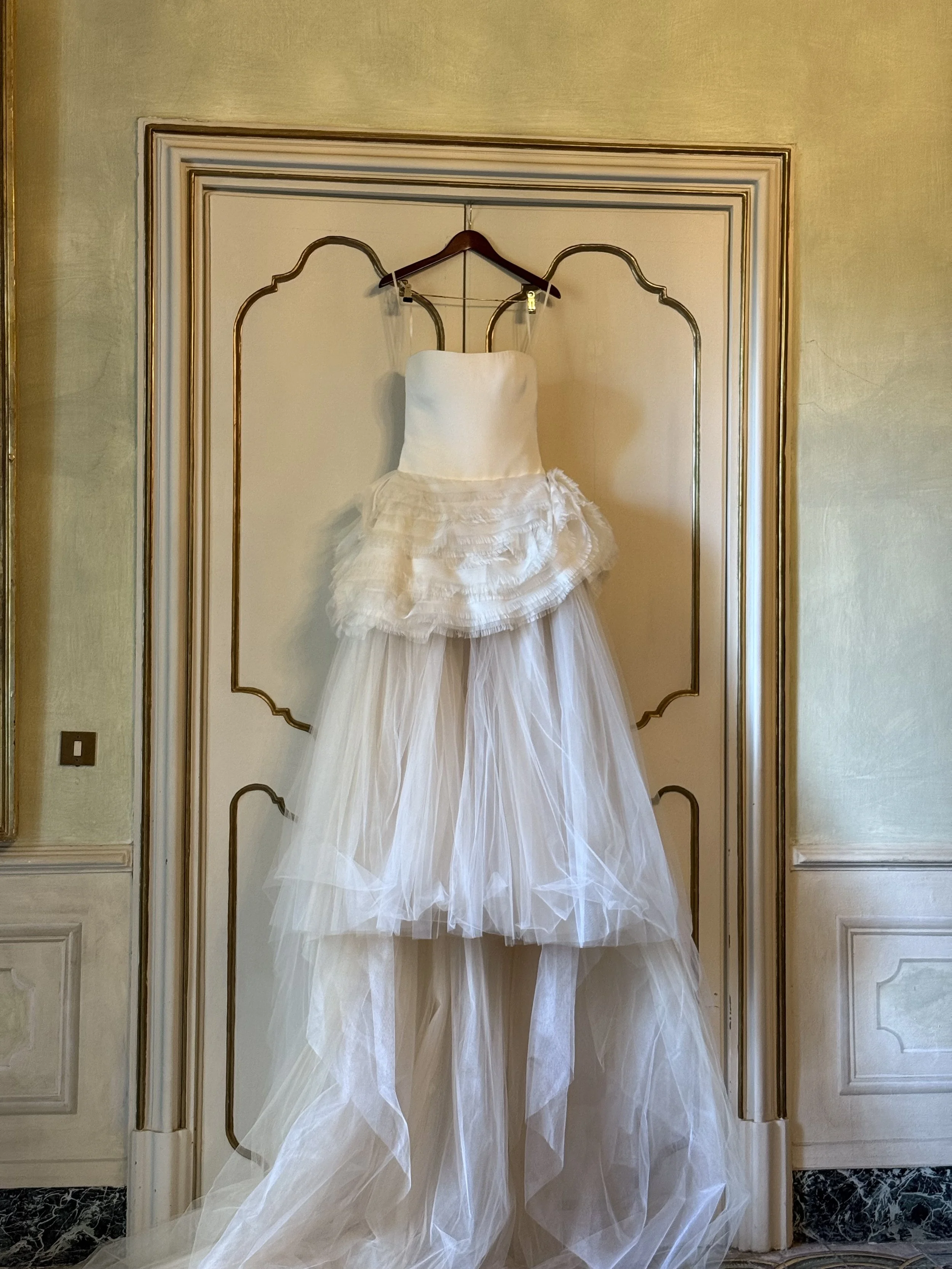 A white wedding dress hanging on a door, featuring a strapless bodice, a layered ruffled midsection, and a long tulle train.