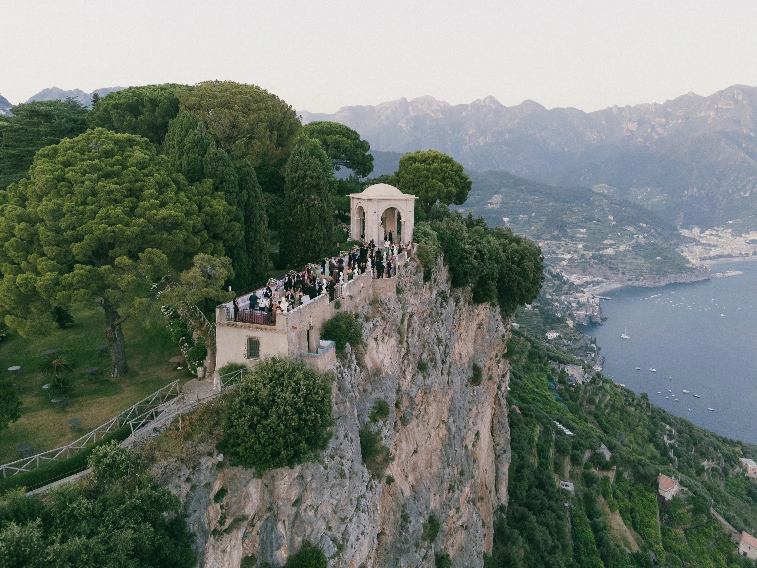 A scenic view of a cliffside garden with a small white pavilion, surrounded by lush green trees, overlooking a lake with boats and a town at the mountain's base. Villa Cimbrone wedding Amalfi Coast