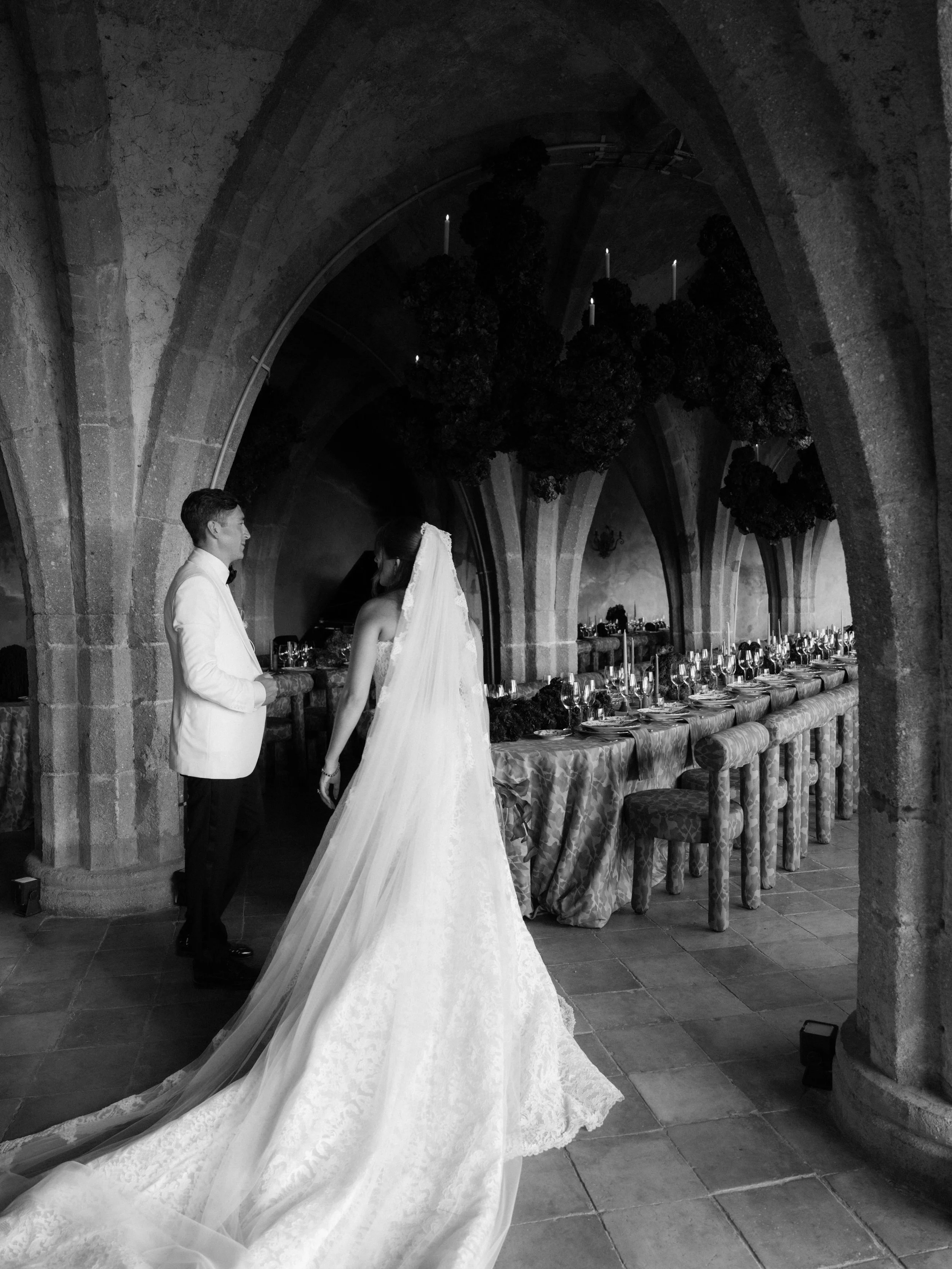 A bride and groom standing face to face in a stone arched hallway during a wedding reception, with a long table set with glassware and floral arrangements behind them. Villa Cimbrone wedding Amalfi Coast