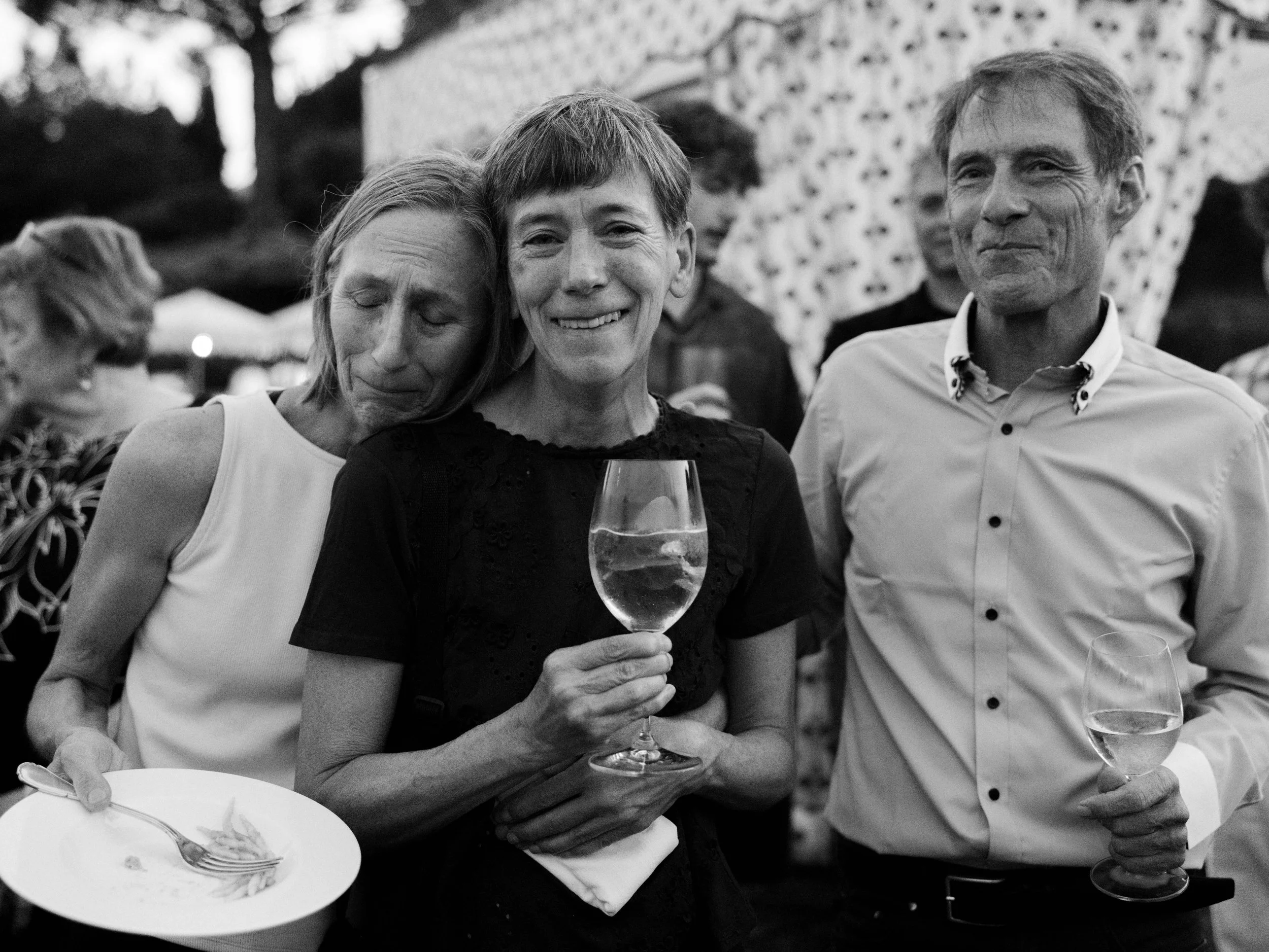 Three people at a social gathering with two women and one man, holding wine glasses, smiling, and hugging each other, with a table and other guests visible in the background.