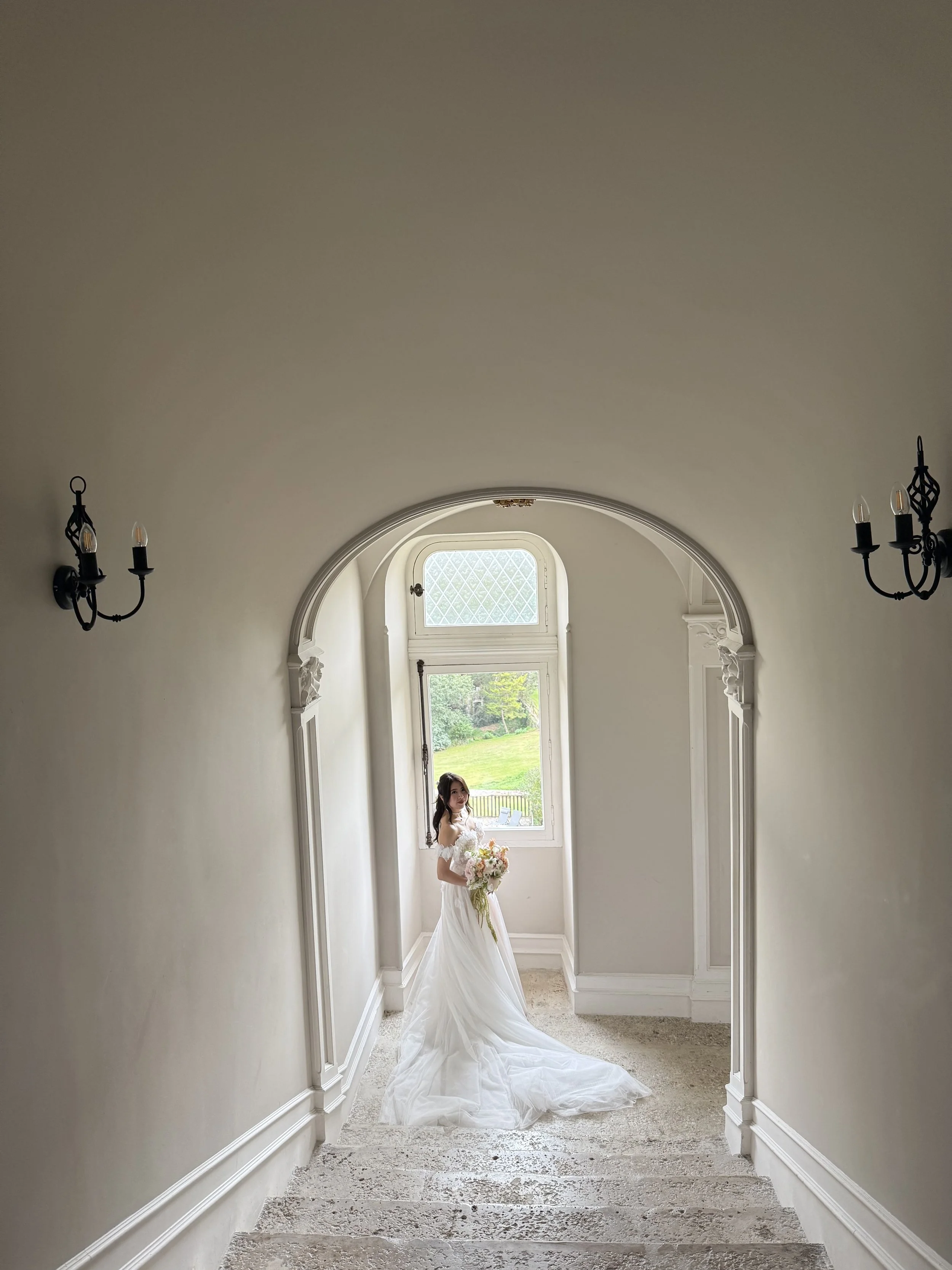 Bride in wedding dress holding a bouquet standing in a staircase landing near a window with lush green scenery outside.