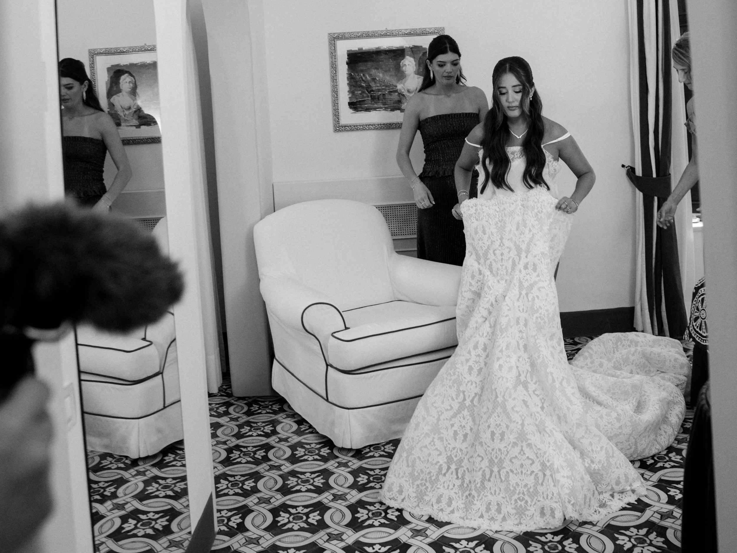 Two women, one sitting and one standing, getting dressed in lace dresses in a room with a patterned floor, a framed painting on the wall, and a mirror reflecting part of the women.