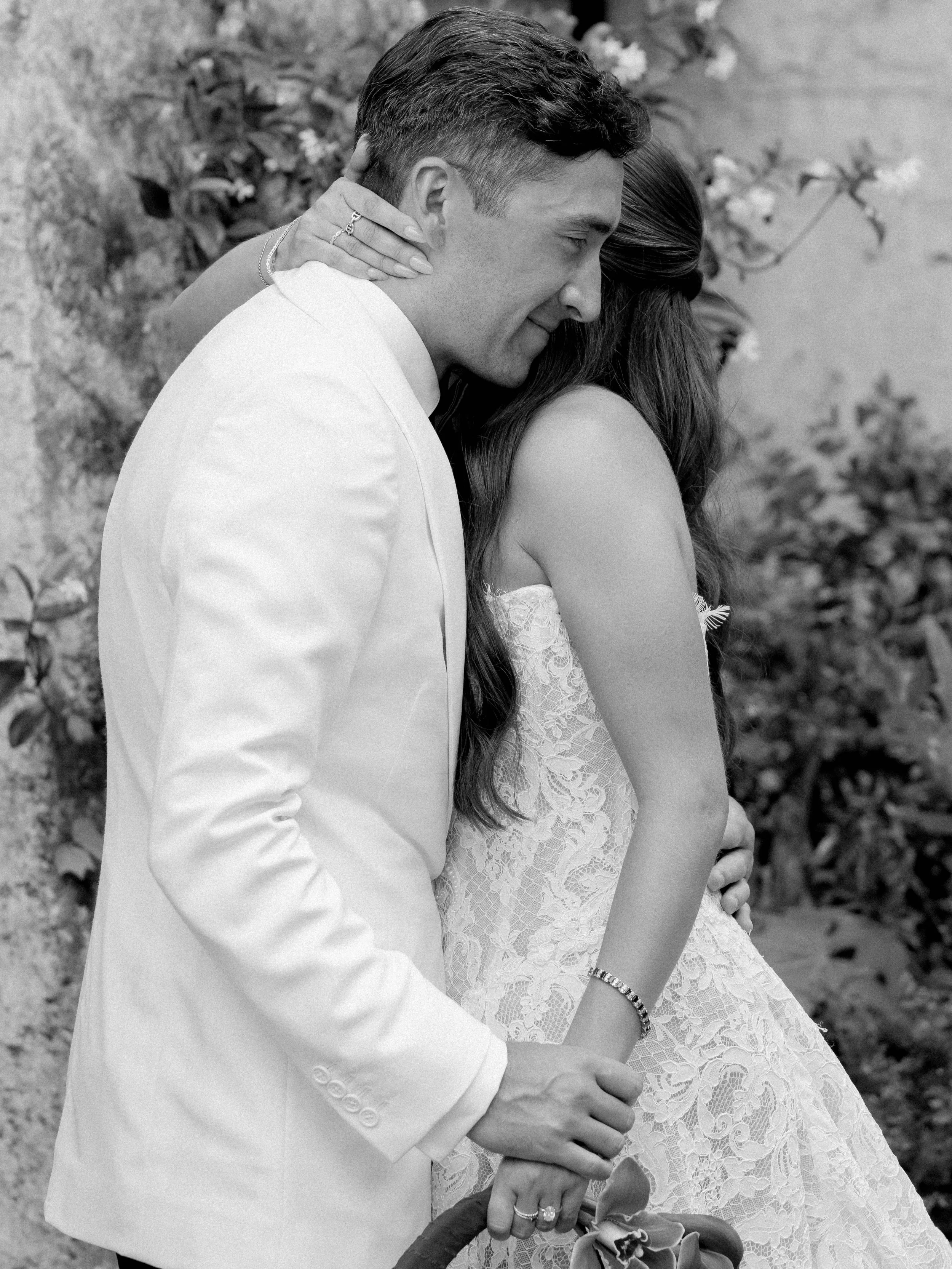 A black and white photo of a couple embracing outdoors, with the man gently holding and kissing the woman's forehead. The woman is wearing a lace dress and the man is in a white suit, both showing affection.