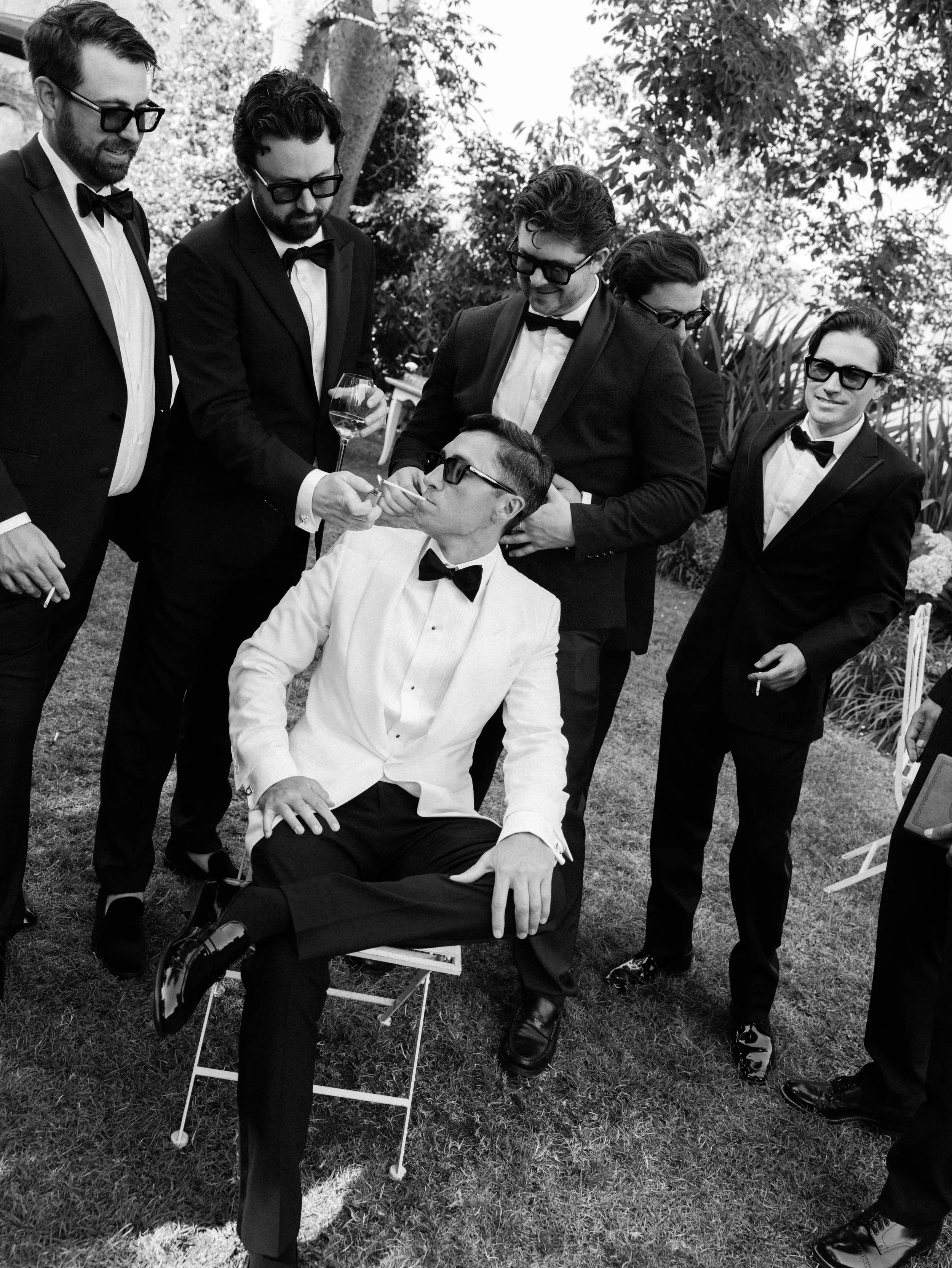 A group of six men dressed in tuxedos and bow ties, some wearing sunglasses, gathered outdoors around a seated man in a tuxedo who is having his face painted or makeup applied, with trees and foliage in the background.