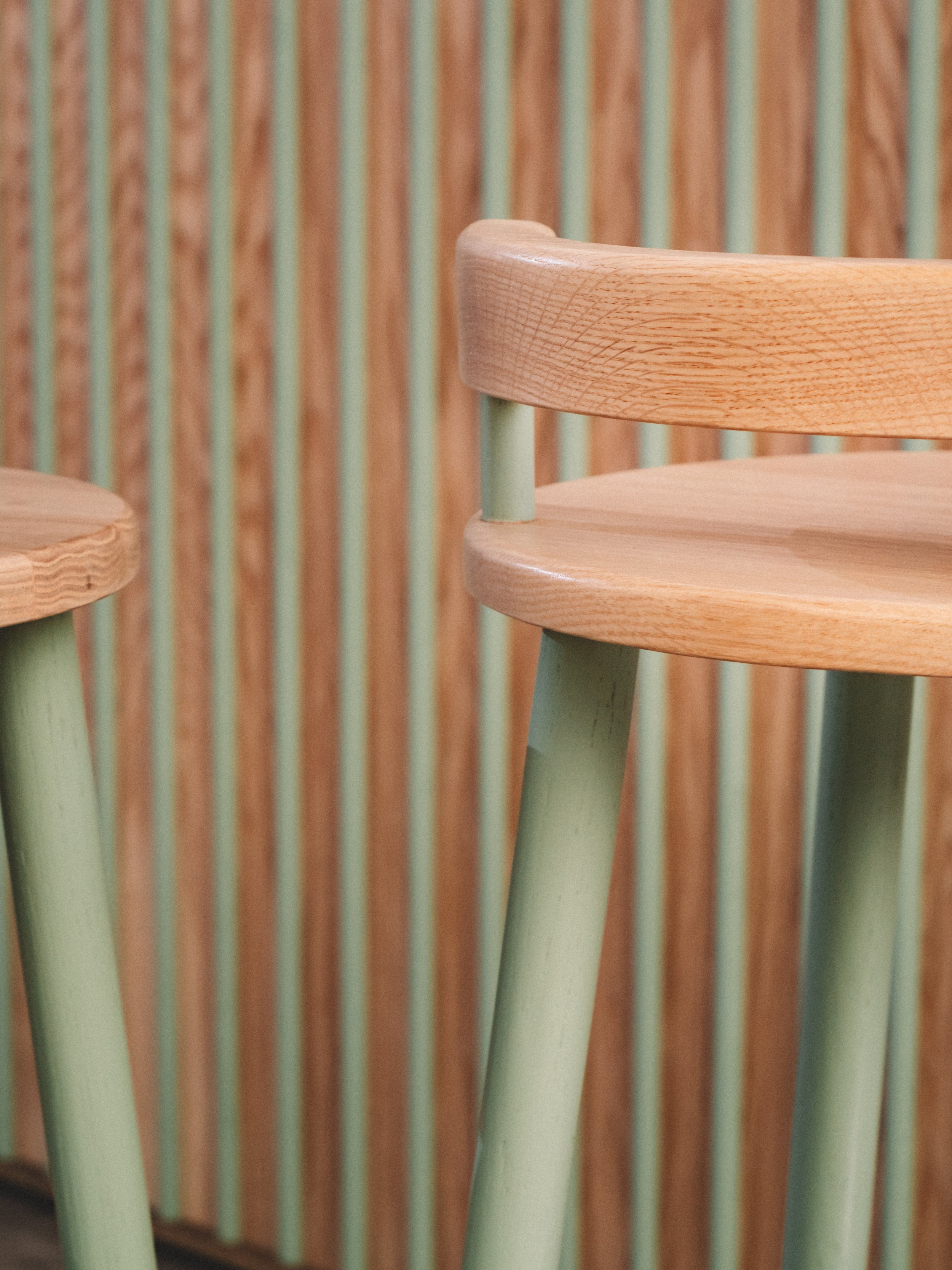 Sillas de madera clara con patas verdes frente a un fondo de paneles de madera y listones verdes.
