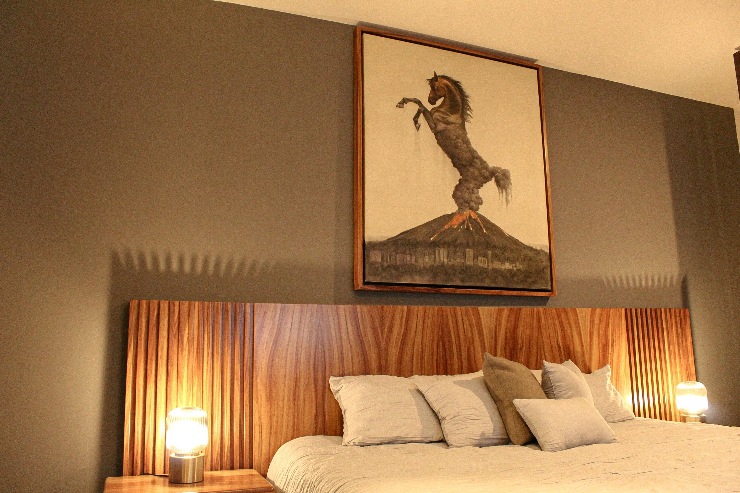 Habitación con cama grande, cabecera de madera, luces en las mesas de noche y cuadro de un caballo saliendo de un volcán.